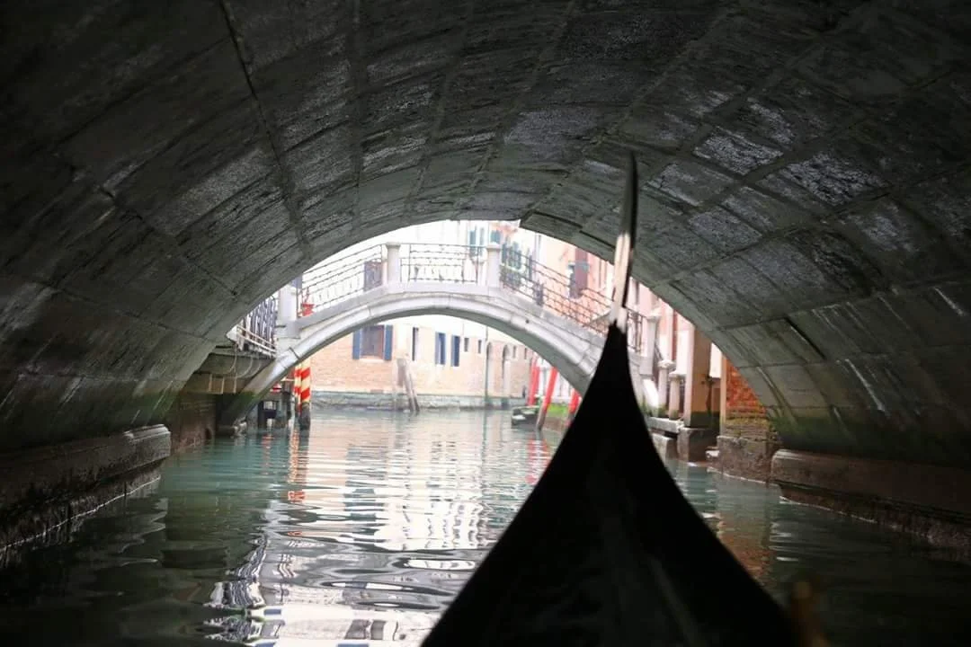 View from inside a gondola under a stone bridge on a canal in Venice, Italy.
