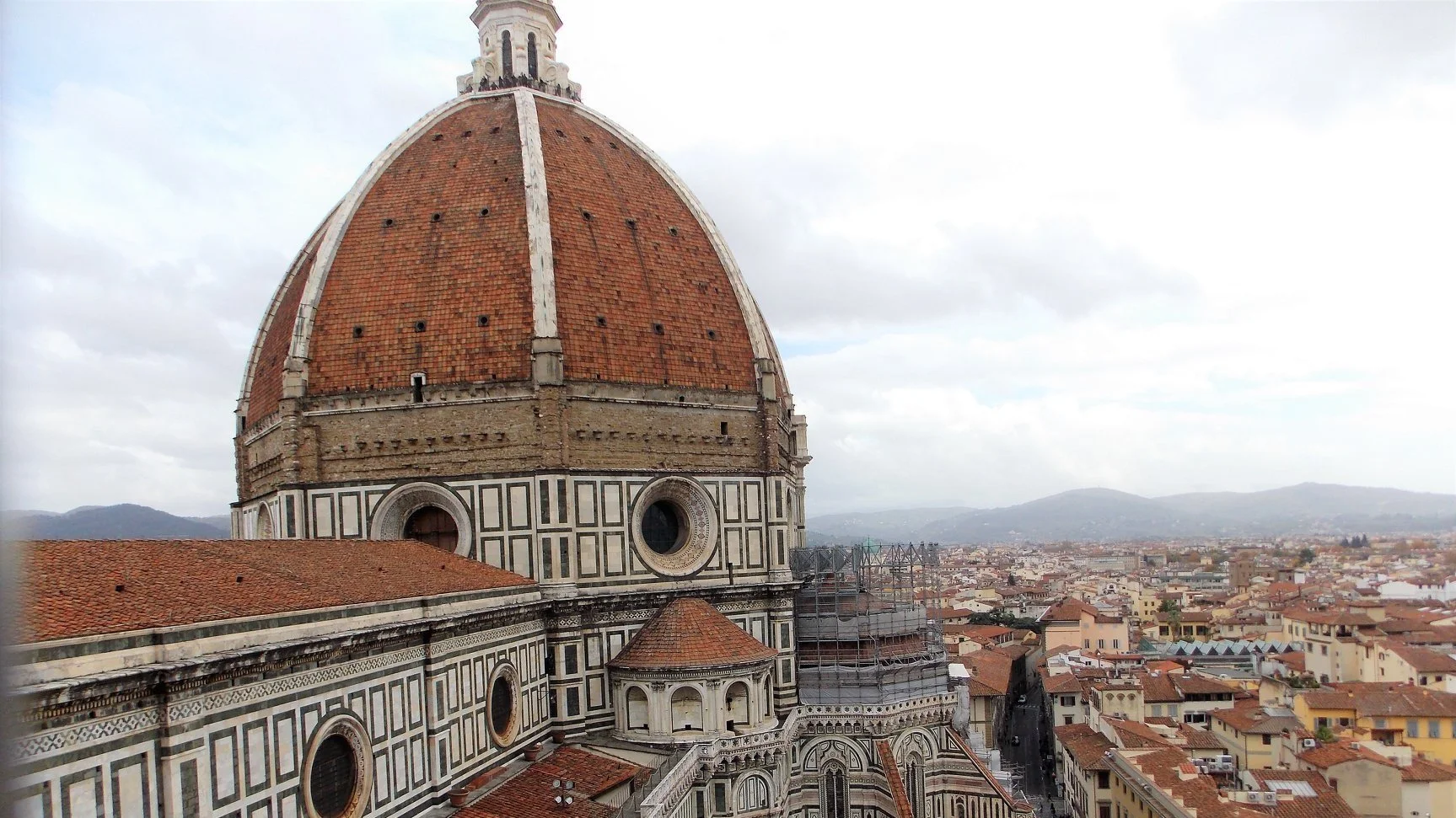 View of the Florence Cathedral’s large terracotta-tiled dome with intricate black and white marble facade, overlooking the city of Florence, Italy, with a cloudy sky in the background.