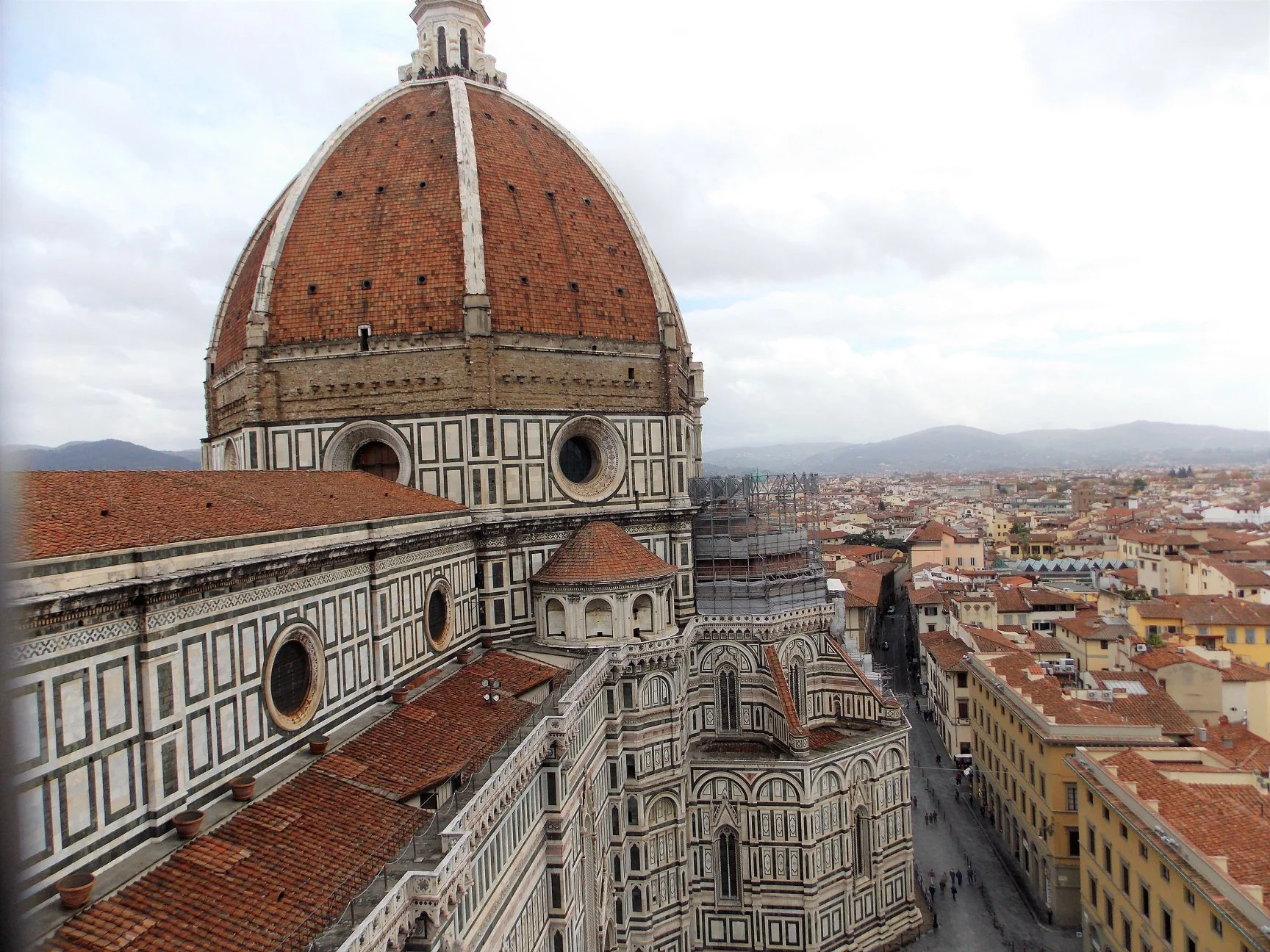 View of the Florence Cathedral, also known as the Cattedrale di Santa Maria del Fiore, with its large red-tiled dome. The cathedral's intricate white, green, and pink marble facade is visible, along with surrounding buildings and a distant cityscape under a partly cloudy sky.