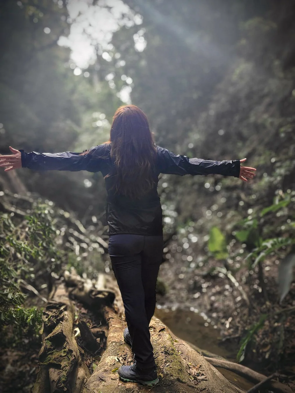 A person with long hair wearing a black jacket and black pants walking on a fallen tree trunk in a forest with arms outstretched.