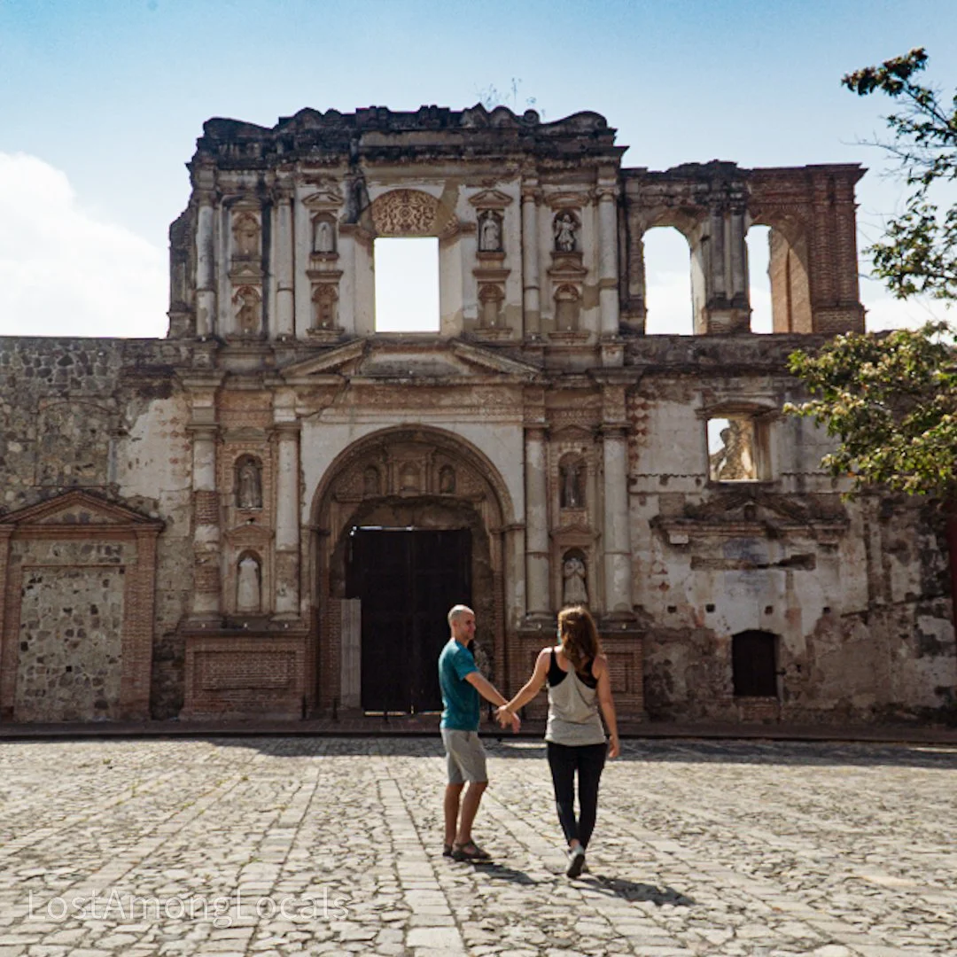 A couple holding hands and walking in front of an ancient, partially ruined stone and brick building with arched doorways and windows, under a clear sky with some clouds.