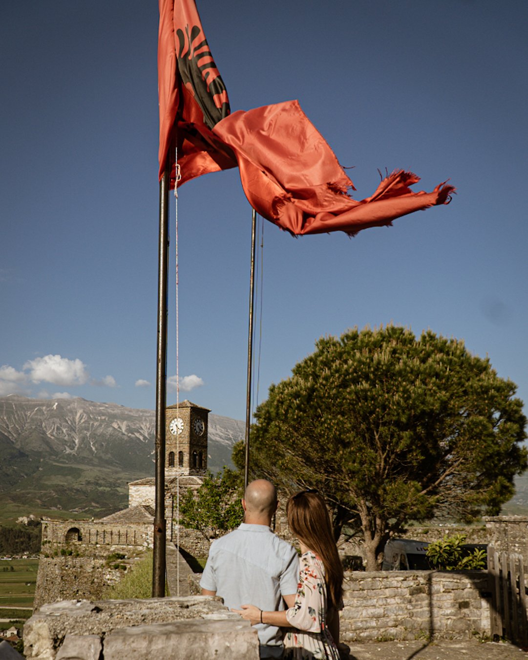 A couple standing together outdoors, facing away from the camera, with a historic stone building, mountains, and a blue sky in the background. Two flagpoles with tattered red flags are in the foreground.