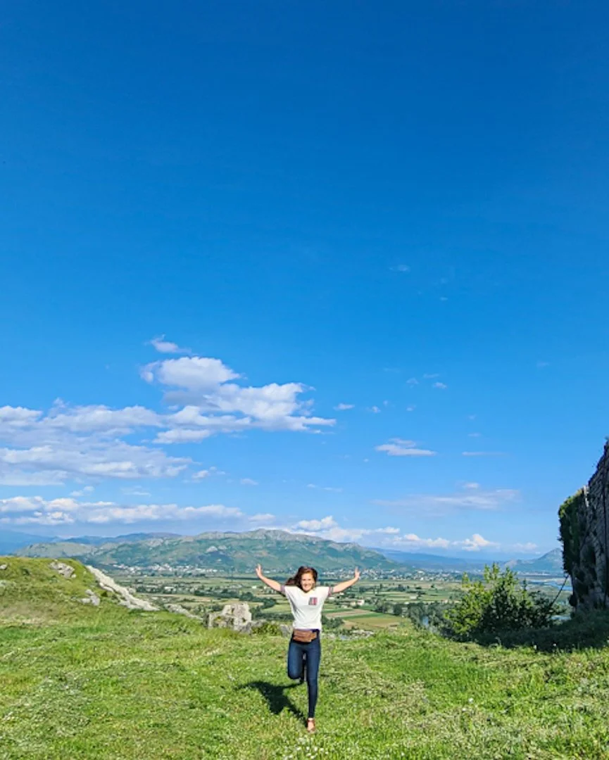Woman in white t-shirt and jeans running on green grass with arms raised, scenic landscape with mountains and blue sky in the background.