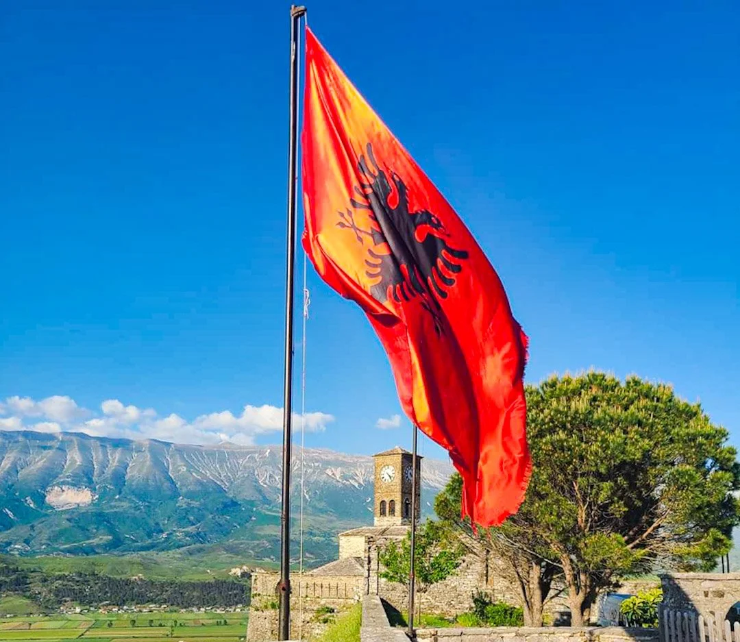 A vibrant Albanian flag with a black double-headed eagle on a red background, flying on a flagpole in front of a historic stone building with a clock tower, lush green trees, and distant mountains under a bright blue sky with a few clouds.