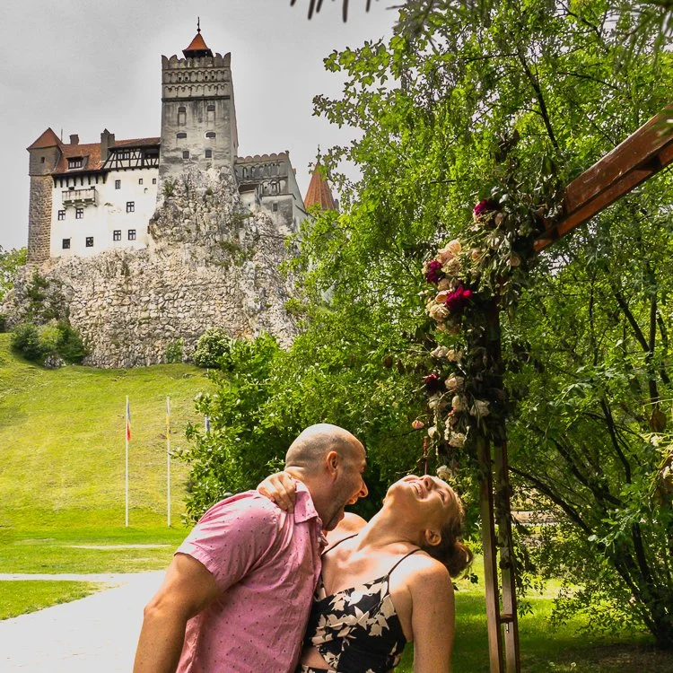 A couple laughing and enjoying each other's company outdoors near a decorated arch, with a castle on a hill in the background.