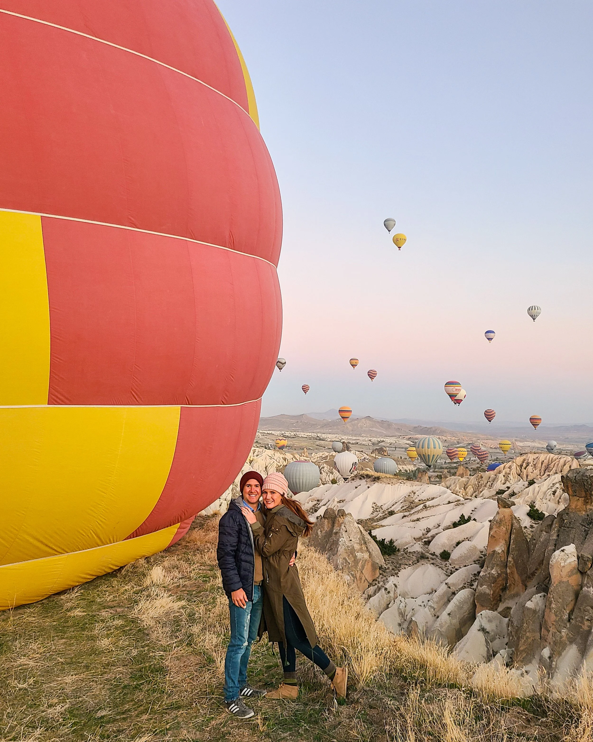 Two women standing in front of a landscape filled with numerous hot air balloons floating in the sky. One woman wears a black jacket and blue jeans, the other wears a green jacket and black pants. They are smiling and hugging each other.