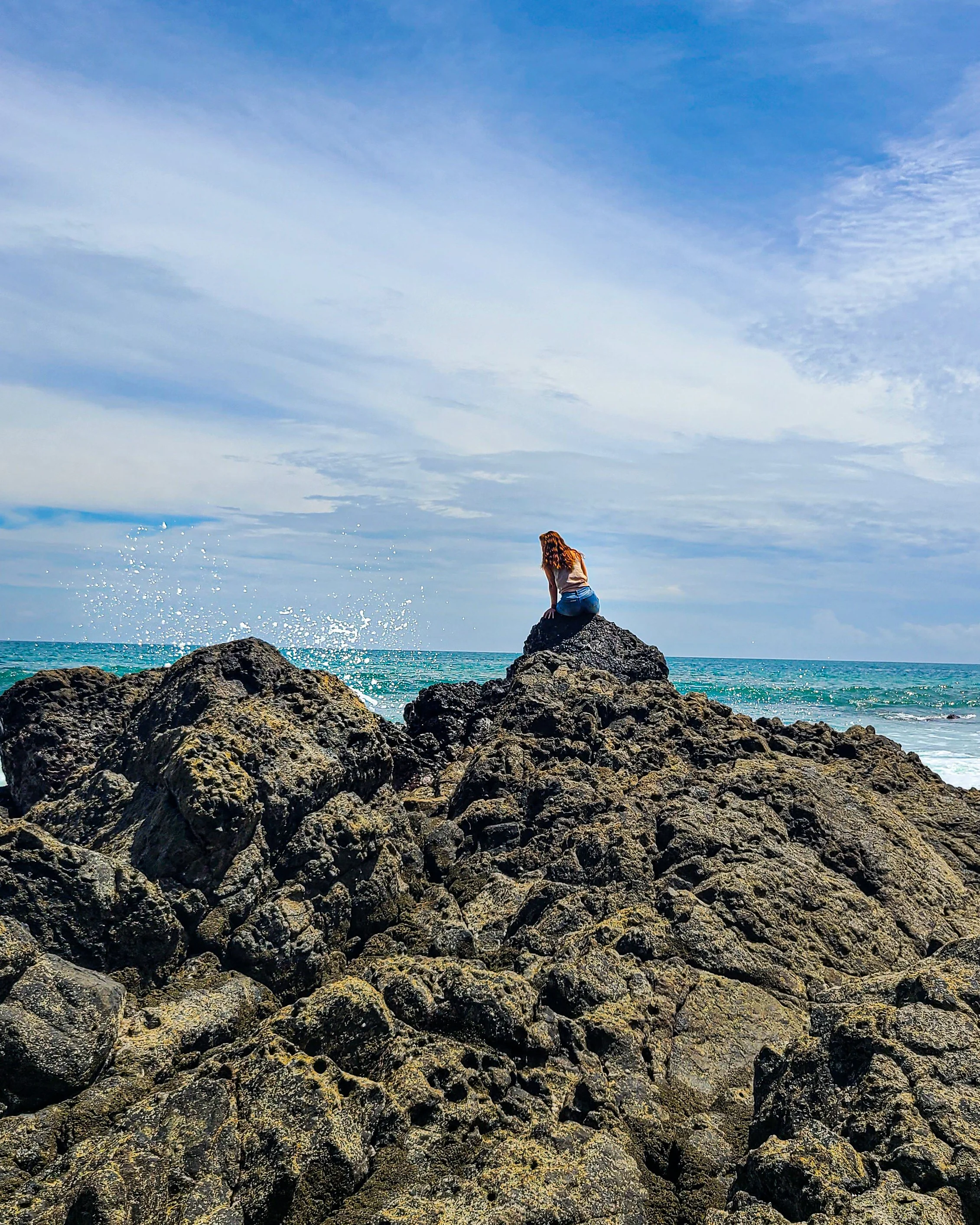 A woman with long hair sitting on rocks by the ocean, looking at the water under a cloudy sky.