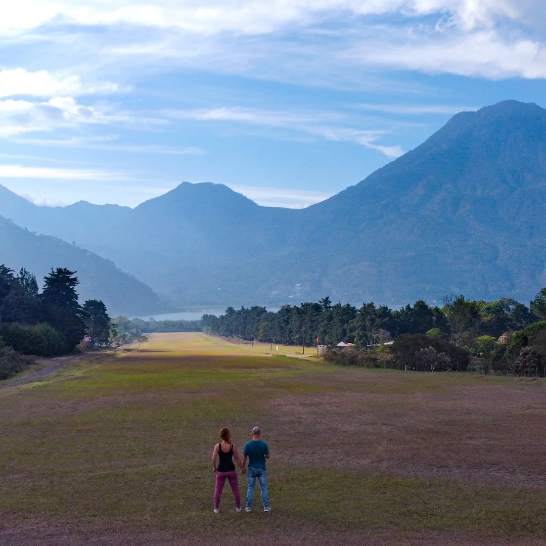 A couple holding hands and gazing at a scenic landscape with mountains, trees, and a cloudy sky.