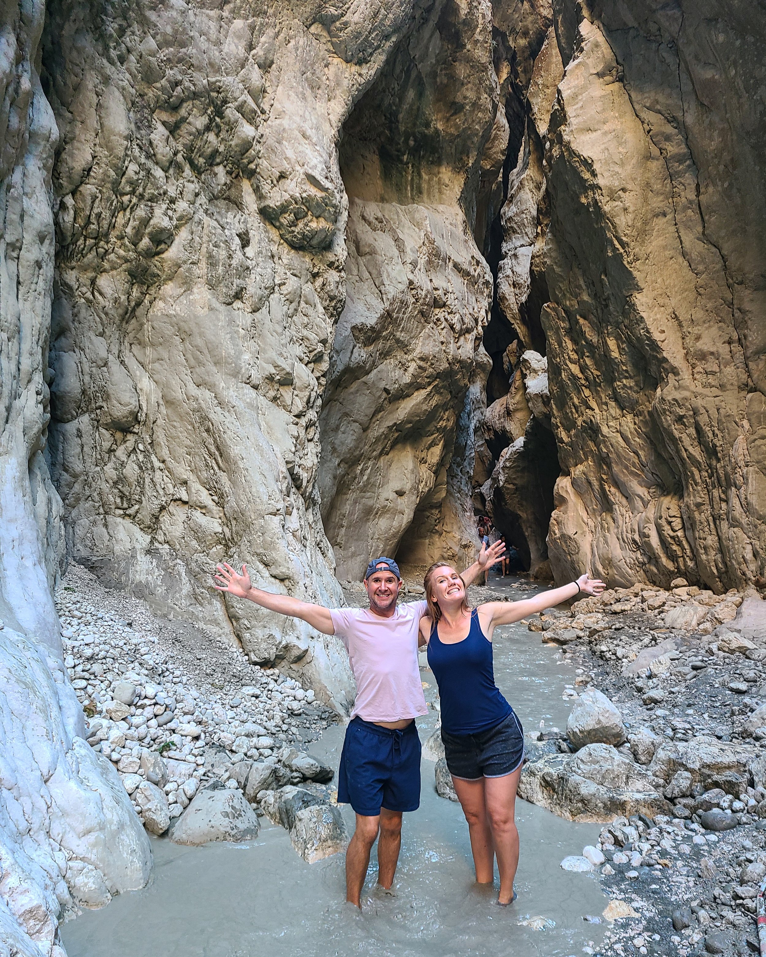 A smiling man and woman with their arms raised, standing in a rocky stream inside a narrow canyon with high, rugged walls.