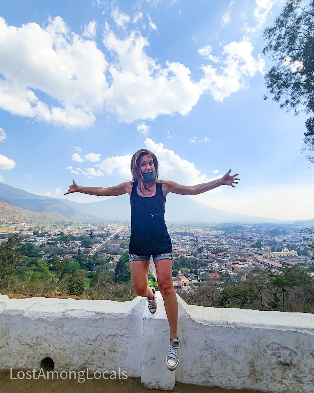 A woman jumping with arms wide open on a white wall, with a cityscape and mountains in the background under a partly cloudy sky.