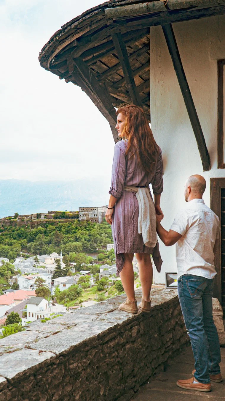 A woman standing on a stone ledge with a man kneeling before her, holding her hand, on a balcony overlooking a town with buildings, trees, and a castle on a hill in the distance during daytime.