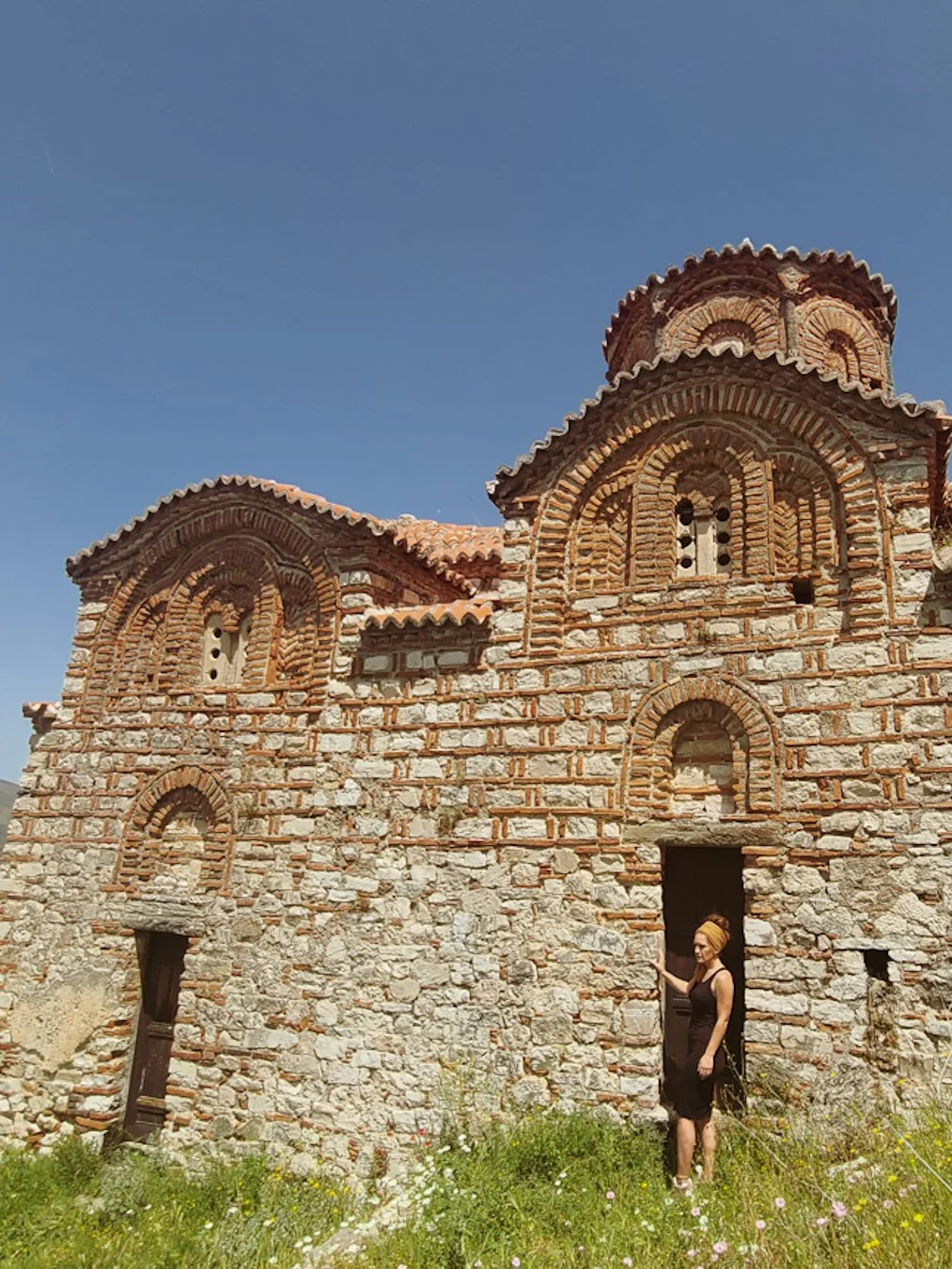 A woman in a black dress and head covering stands outside a small, ancient stone church with brick arches and small windows, under a clear blue sky.