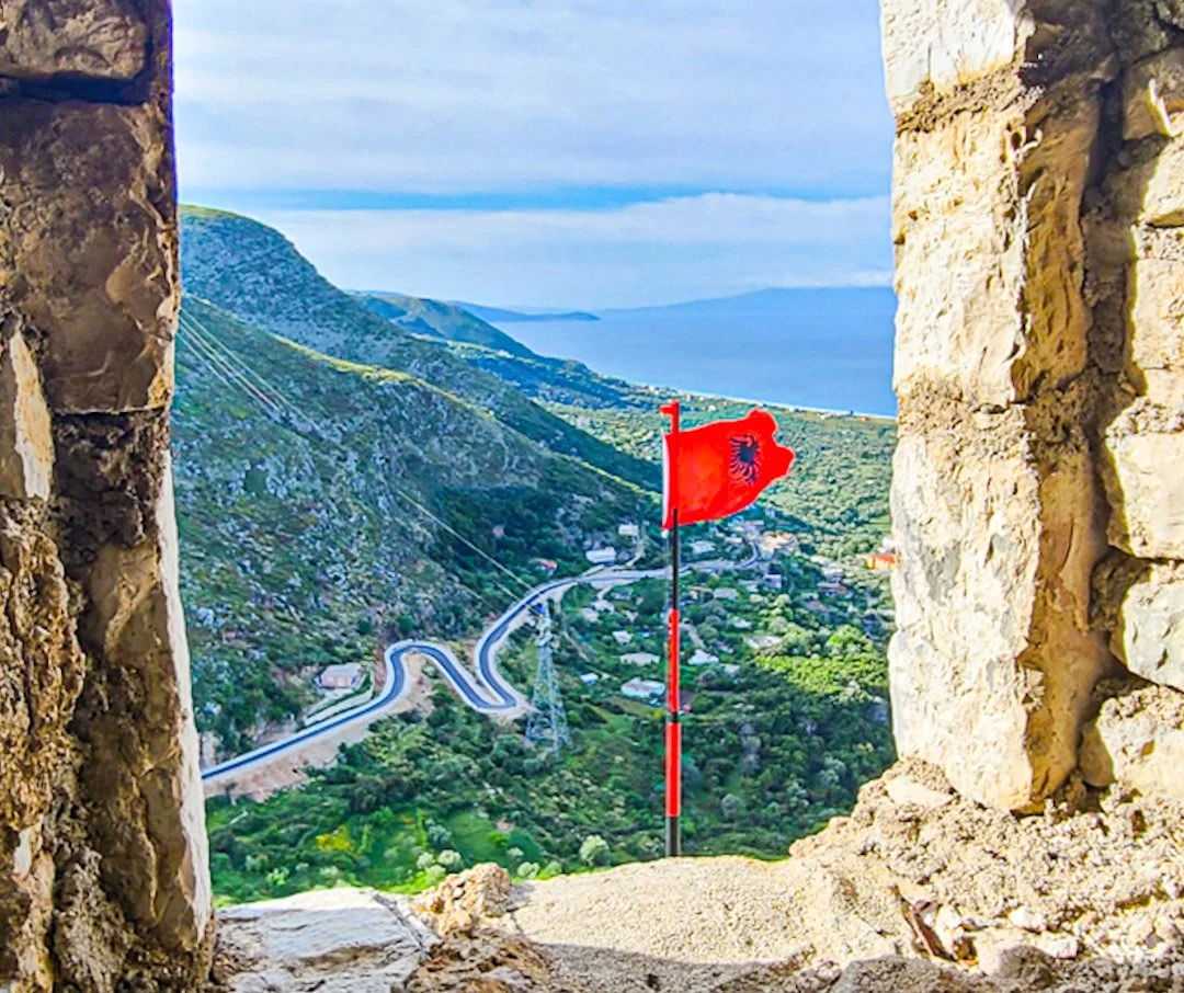 View of a scenic landscape with winding road, green hills, and the ocean in the background, seen through an opening in a stone wall; a red flag with black emblem on a pole in the foreground.