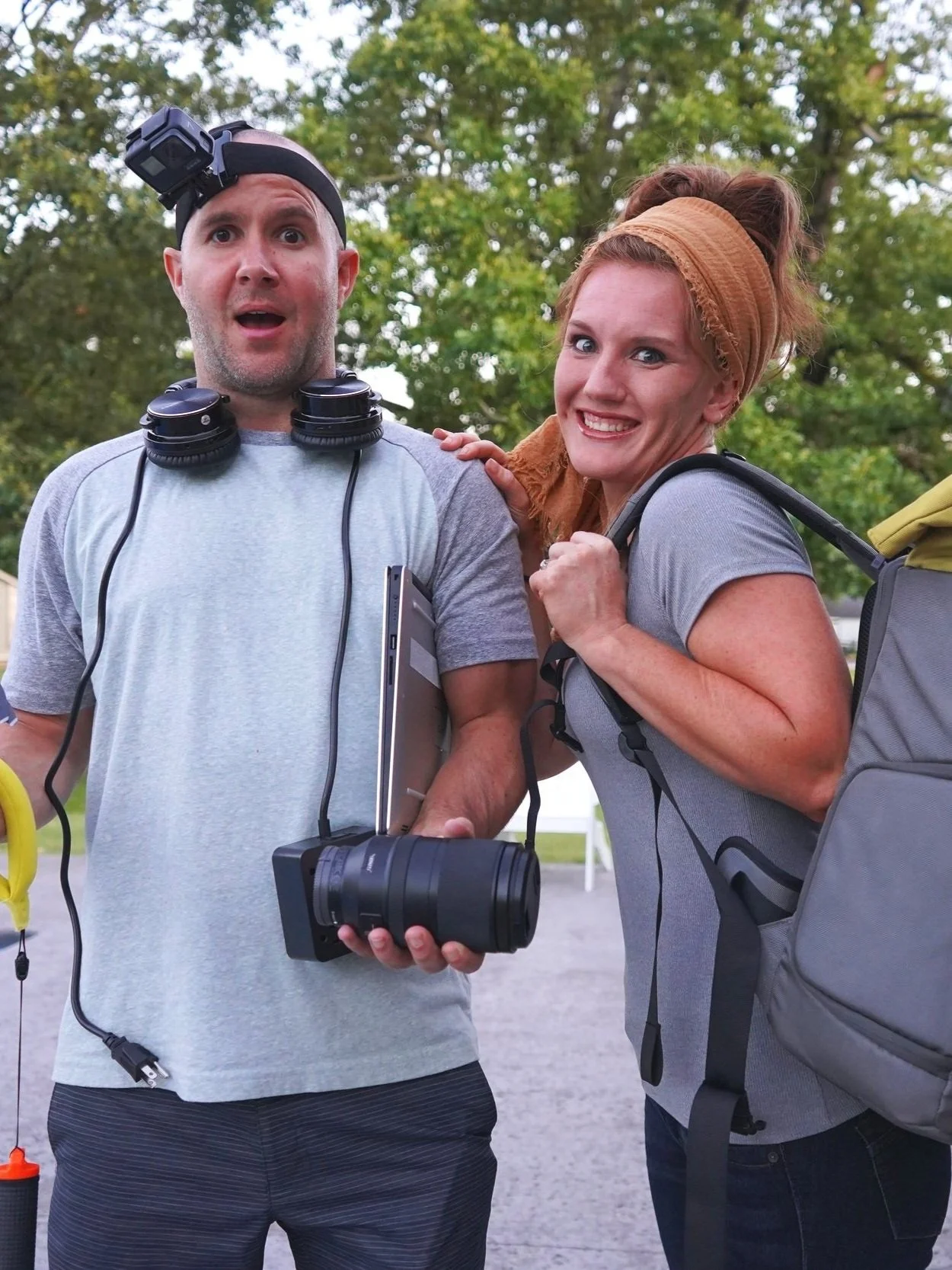 A man and woman outdoors with trees in the background. The man wears a headlamp, headphones, and holds a camera with a large telephoto lens. The woman has red hair in a bun, wears a headscarf, backpack, and gray shirt, and is smiling at the camera.