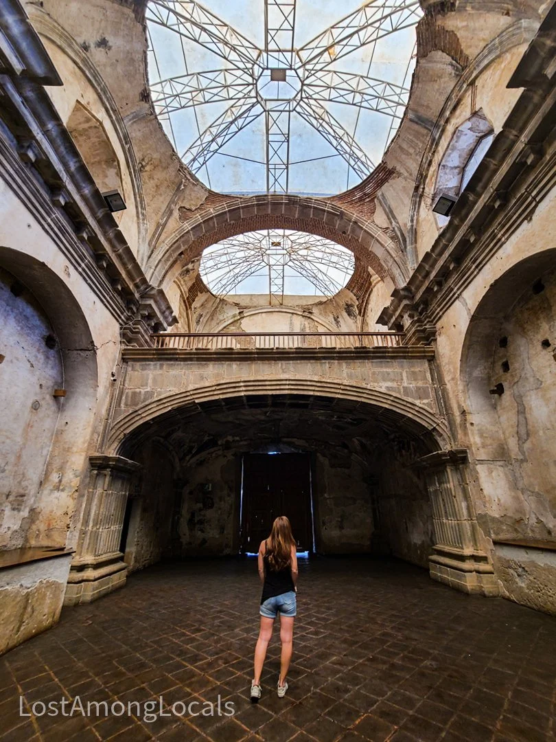 A woman stands inside an old, abandoned building with a high, open ceiling that reveals a metal framework and a partially damaged dome. The interior walls are aged and weathered with arches and columns, and the floor is made of dark bricks.