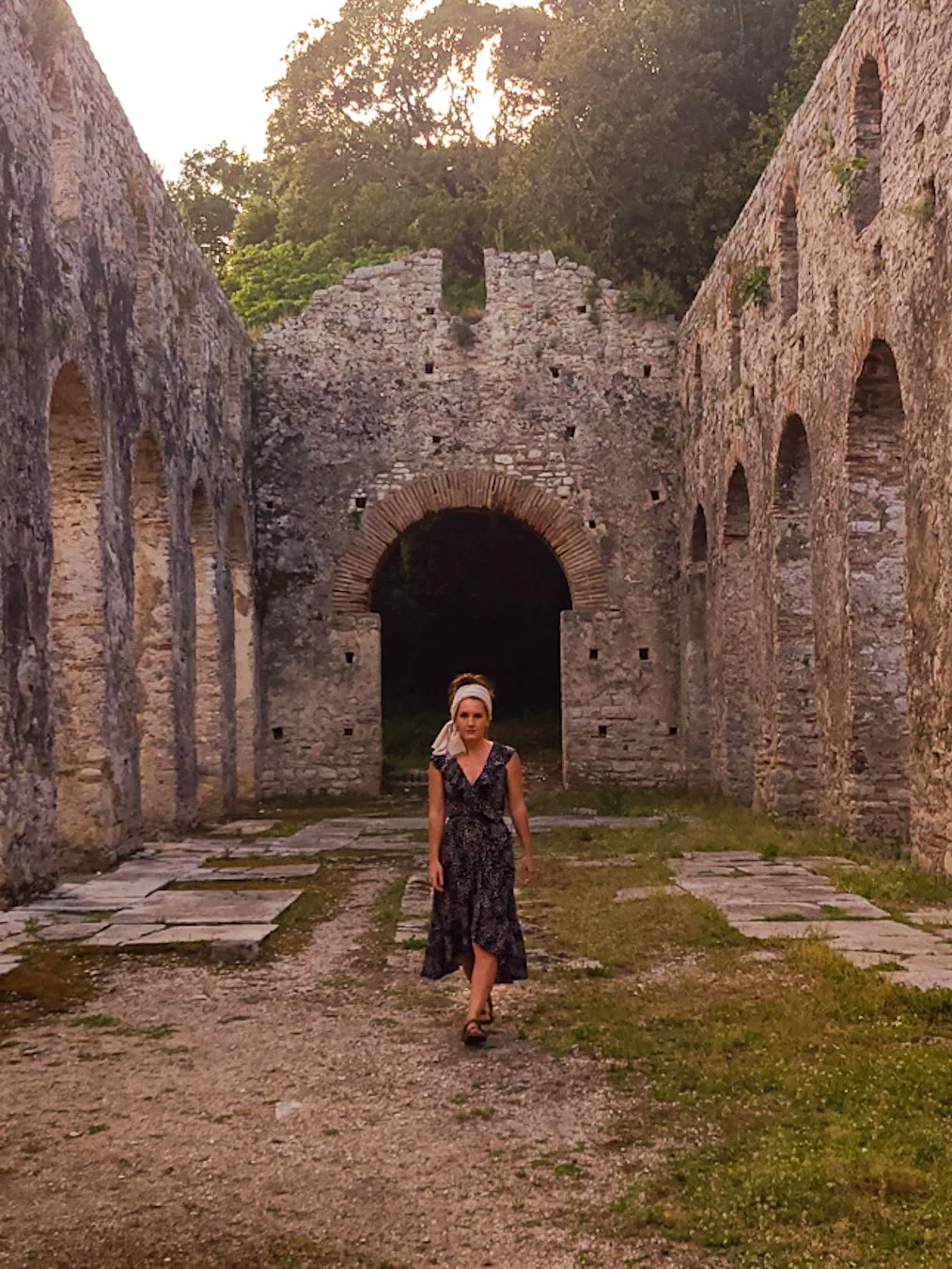 A woman wearing a dark floral dress and a headscarf walking through the ruins of an ancient stone structure, with large archways and tall stone walls, during sunset.