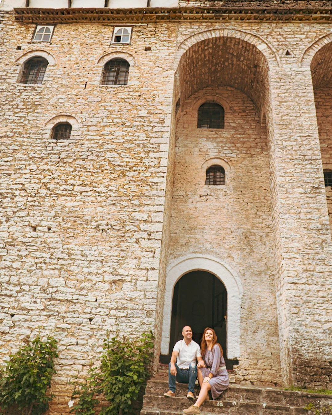 A couple sitting on the steps in front of an old stone building or castle with small windows and an arched entrance, smiling and holding hands.