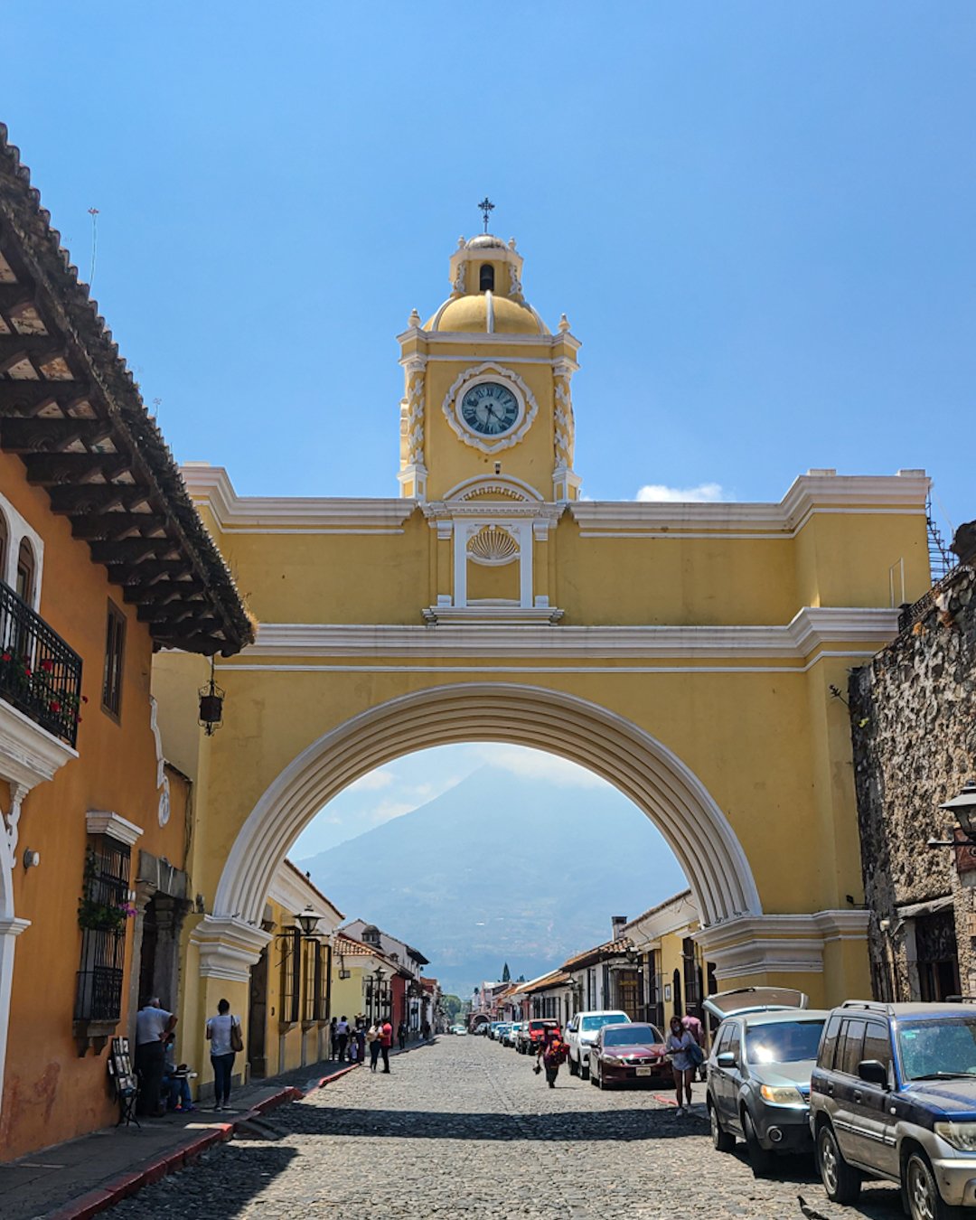 A yellow archway with a clock tower on top in a historic town street, people walking, and cars parked along cobblestone road, with a mountain visible in the distance.