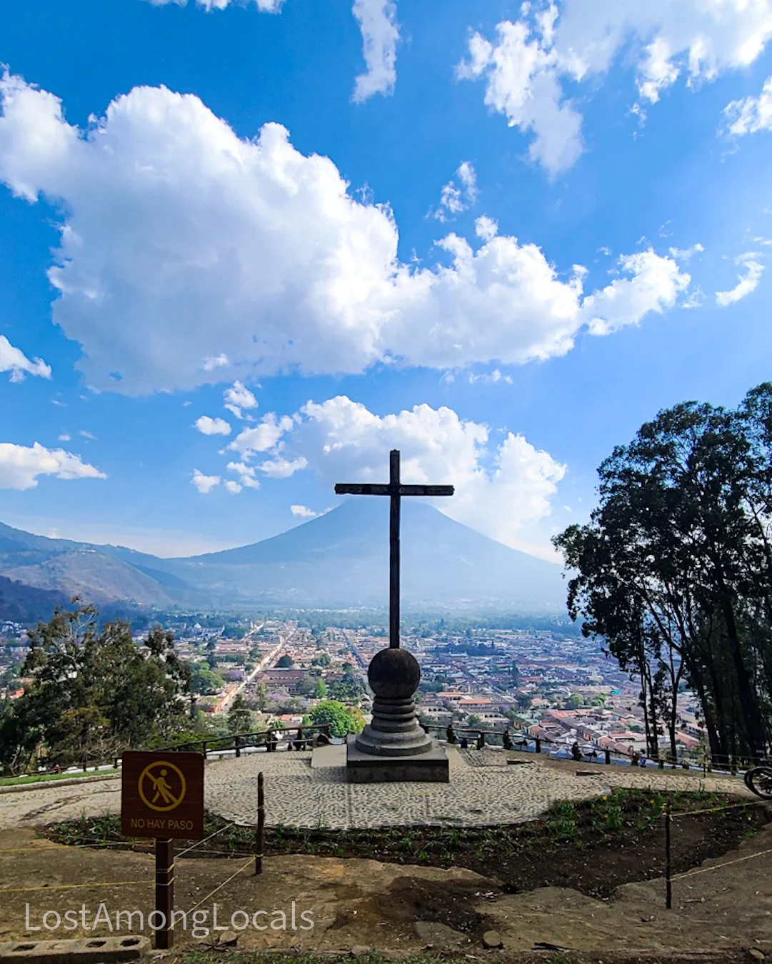 A hilltop with a large cross monument overlooking a city, with mountains and a blue sky with clouds in the background.