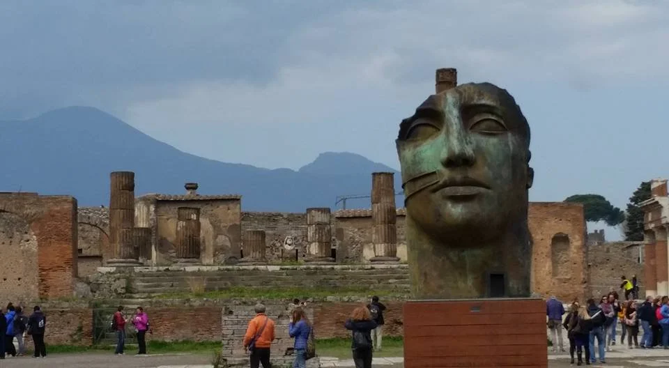 A large sculpture of a human head in front of ancient ruins with columns and steps, and a mountain in the background, at an archaeological site.