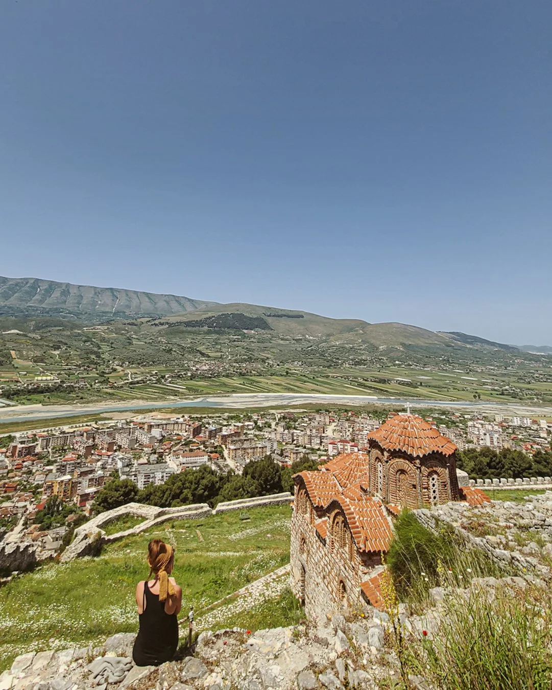 A woman sitting on a grassy hill, overlooking a historic church with red-tiled roof and a town below, with mountains and a river in the background.