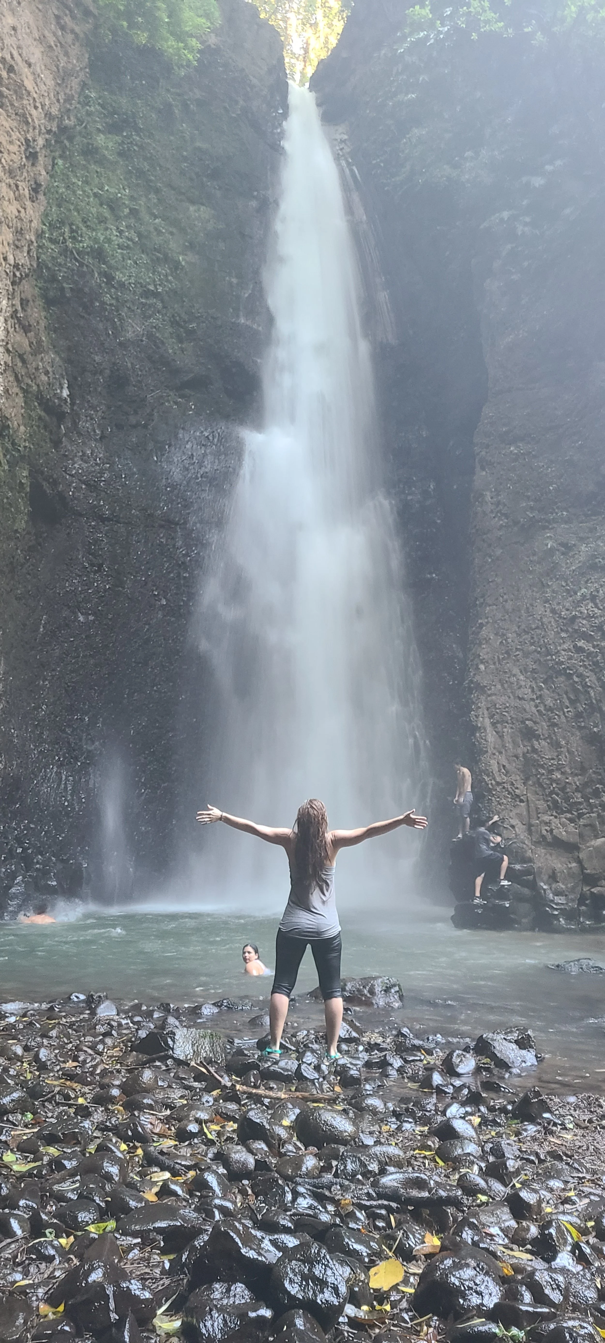 Person standing with arms outstretched in front of a tall waterfall in a rocky, forested area.