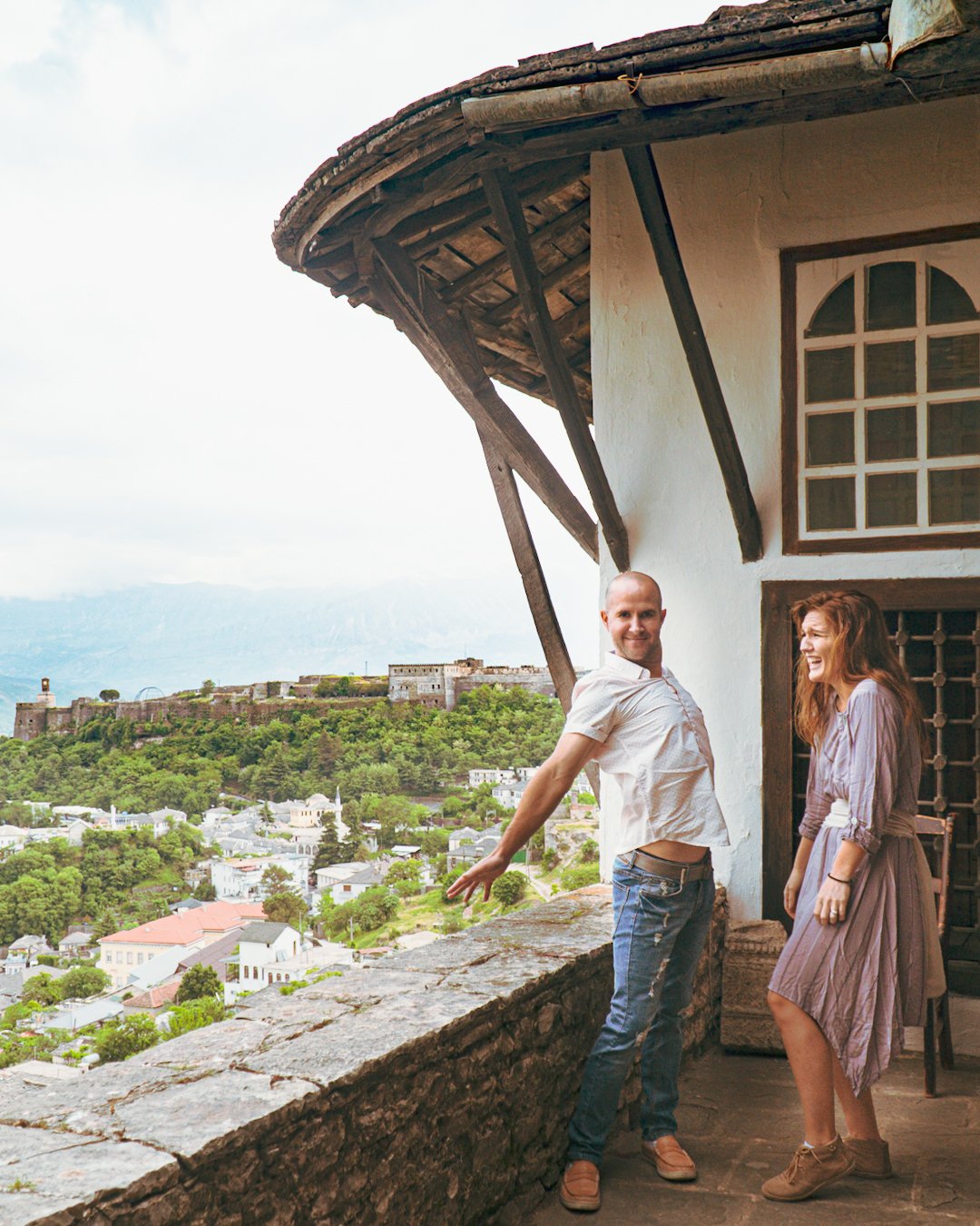 Two people standing on a balcony of a historic building with a scenic view of a city, greenery, and a fortress on a hill in the background.