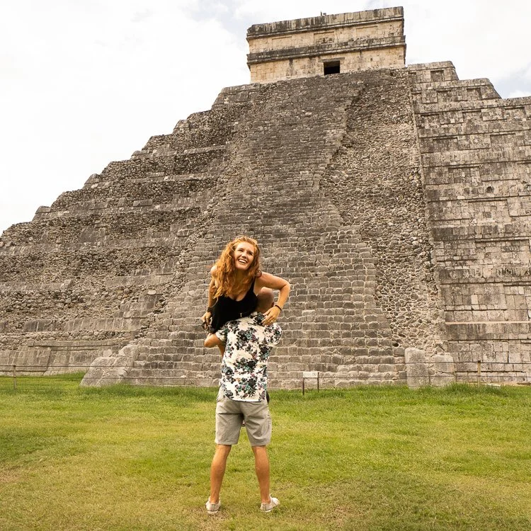 A smiling woman with curly red hair sitting on a man's shoulders in front of a large Mayan pyramid at Chichen Itza in Mexico, with green grass in the foreground and a cloudy sky above.
