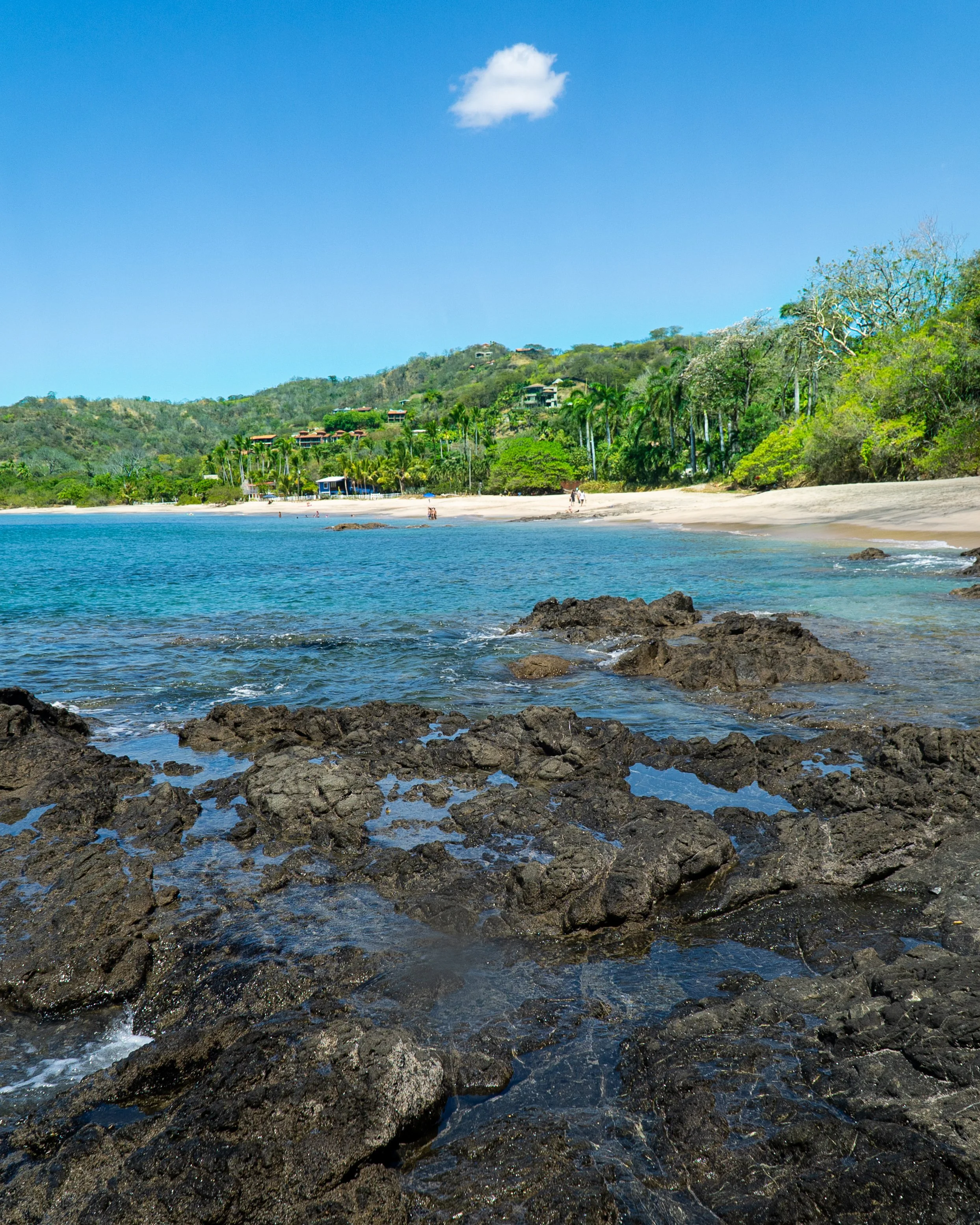 A scenic beach with dark volcanic rocks in the foreground, calm blue water, a sandy shoreline with a few people, lush green trees and hillside with rooftops and houses, and a single fluffy white cloud in the clear blue sky.