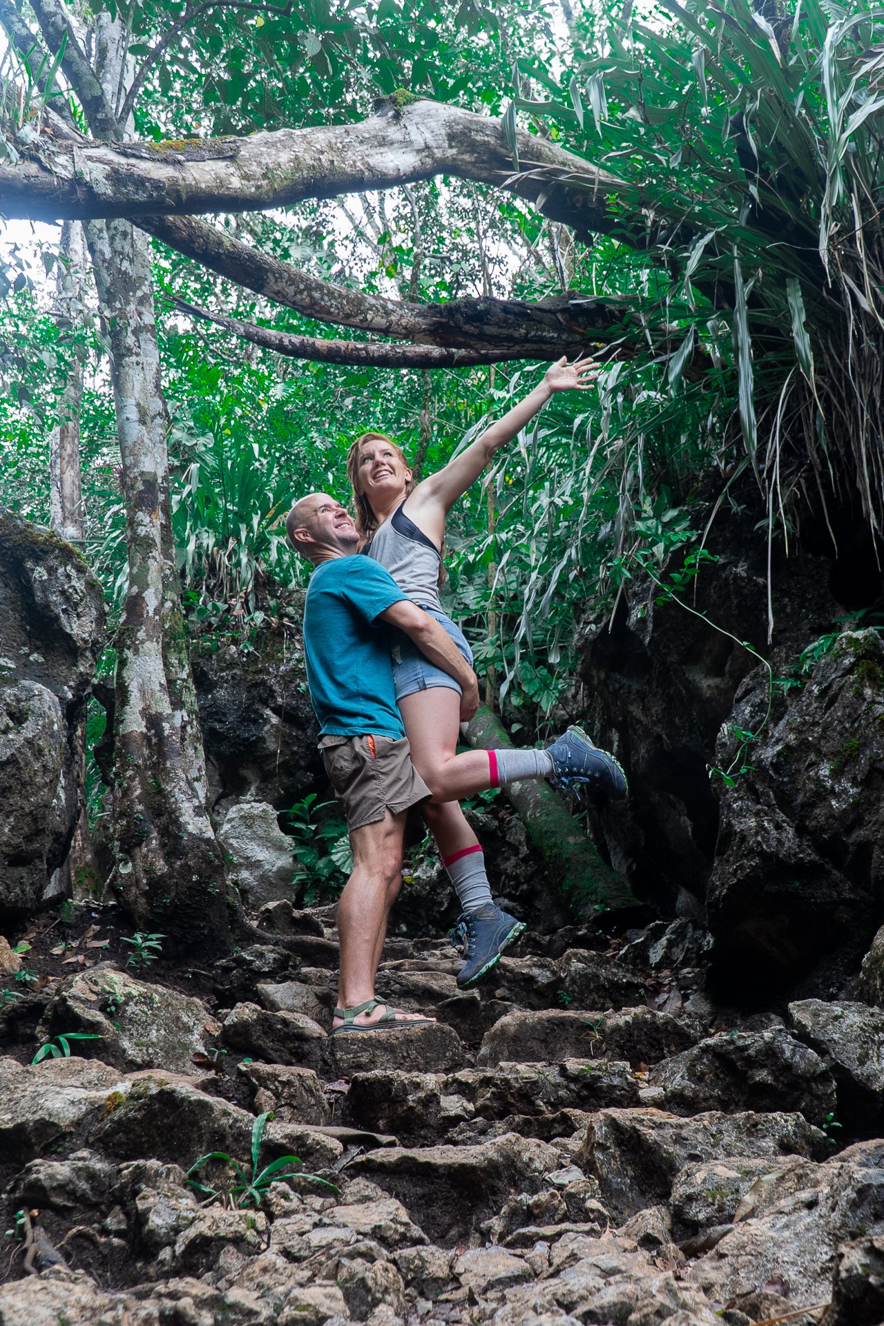A man is holding a woman in a hiking trail with rocks and surrounded by dense green forest, with the woman reaching toward a tree branch.