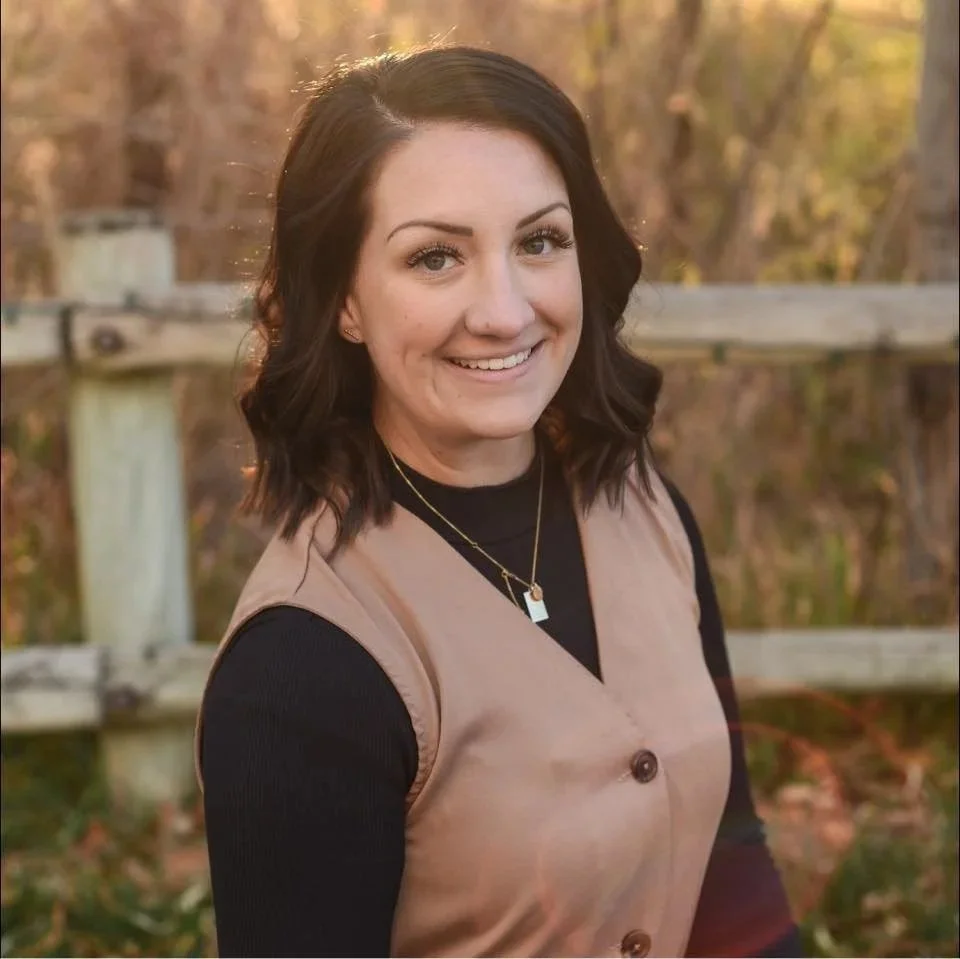 A woman with dark wavy hair smiling outdoors during autumn, wearing a beige vest over a black top, with trees and a wooden fence in the background.