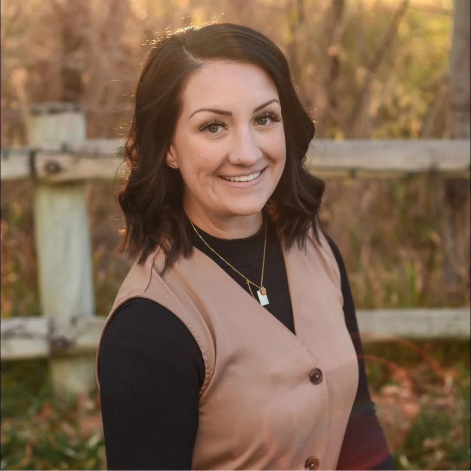 A woman with dark brown hair, smiling, standing outdoors near a wooden fence, wearing a black long sleeve shirt and a tan sleeveless vest with large buttons, and gold necklaces.