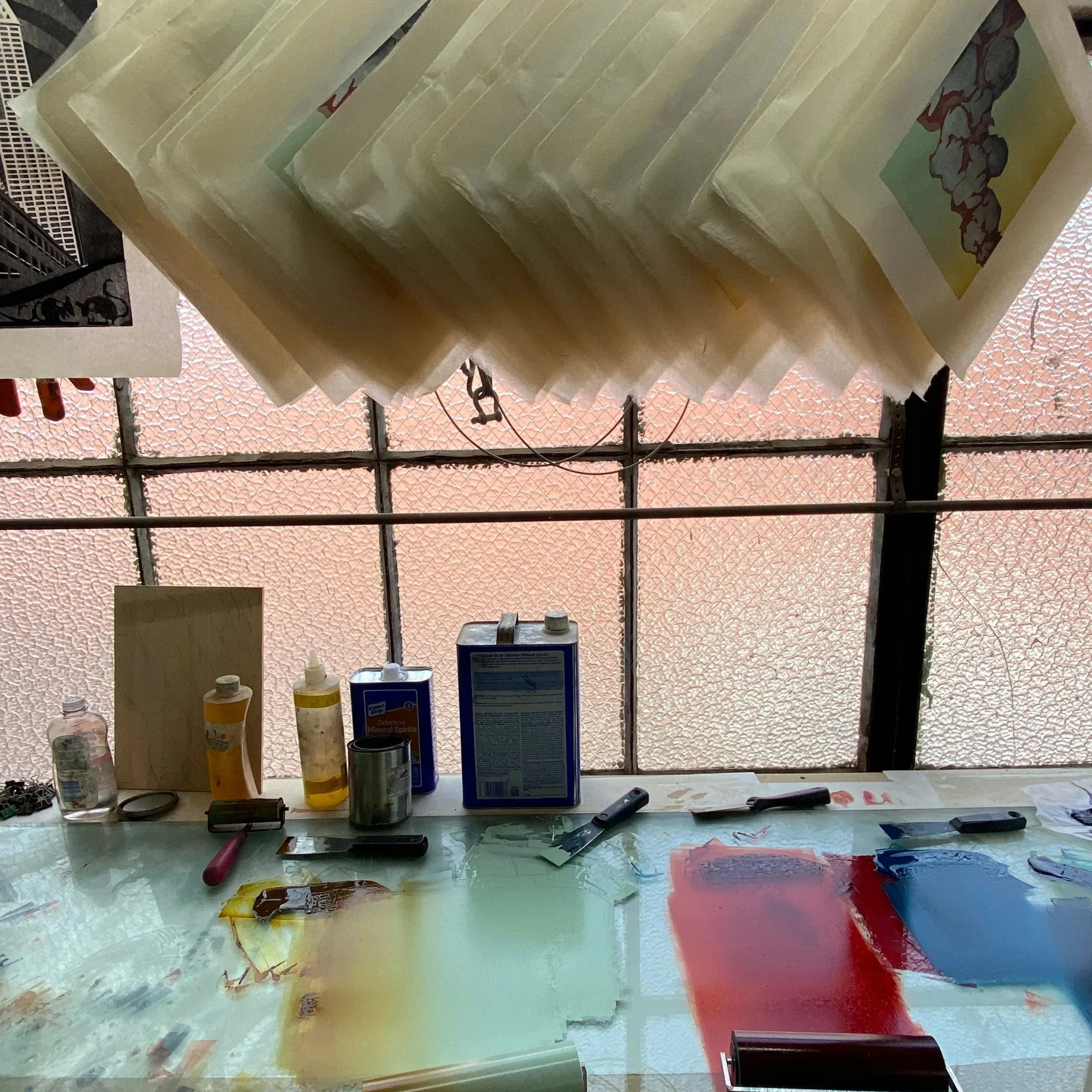 Art studio workspace with paint supplies, brushes, and painted samples on a table, with textured glass window in the background.