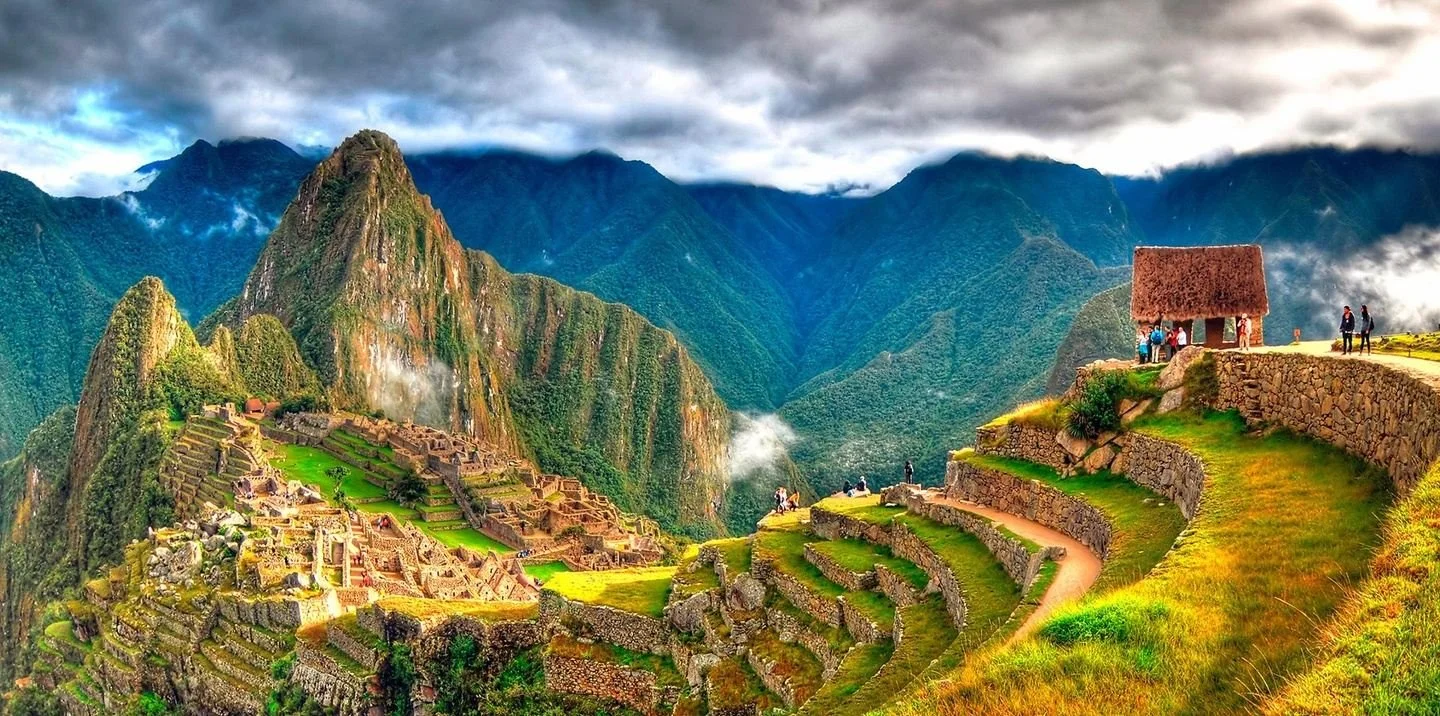 Terraced mountainside with ancient Incan ruins and a small thatched hut, overlooking lush green valleys and a cloudy mountain range in the background.