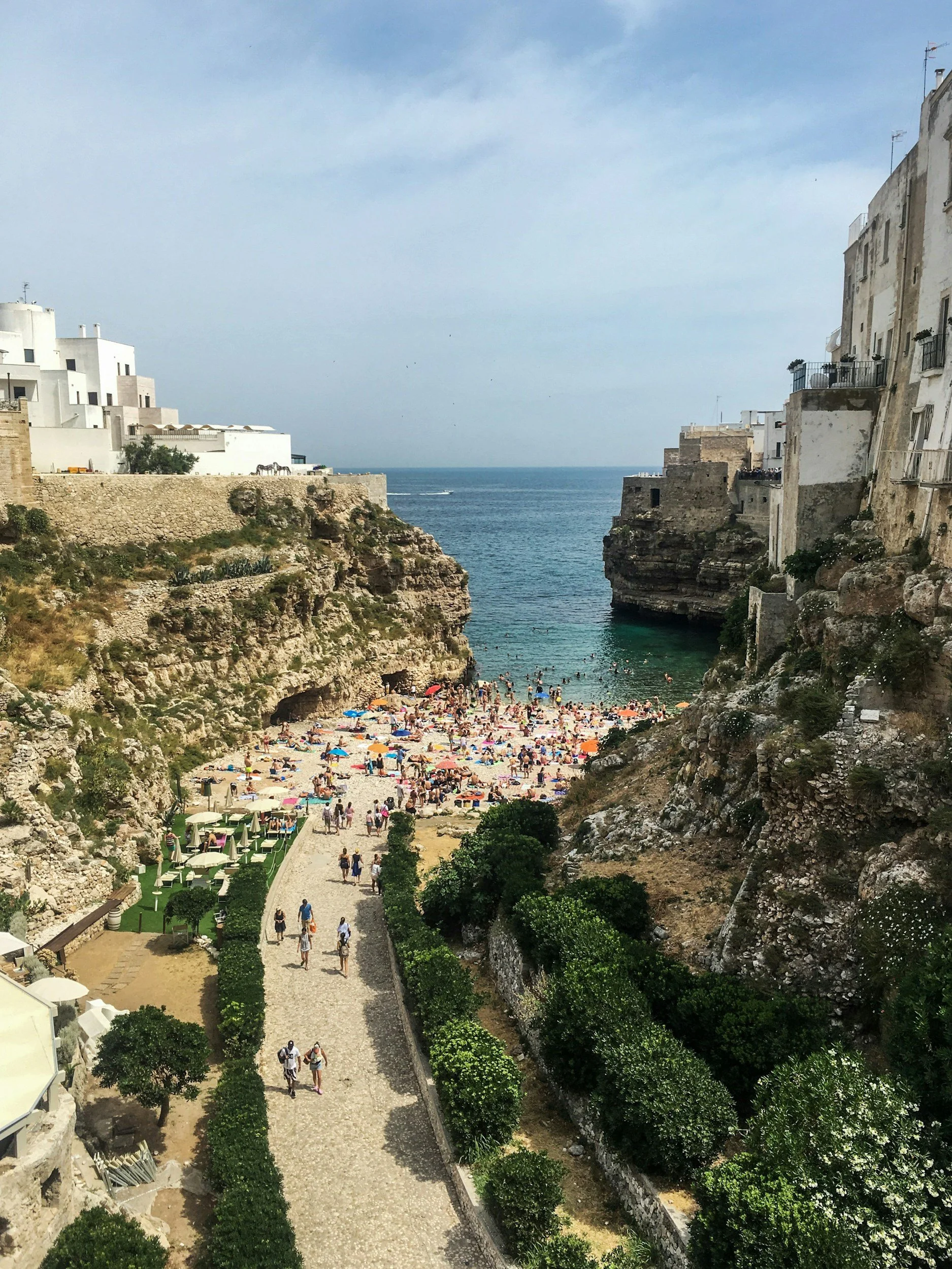 A beach scene in a cove with cliffs on either side, crowded with people sunbathing, swimming, and relaxing under umbrellas. Buildings are visible on the cliffs overlooking the beach.