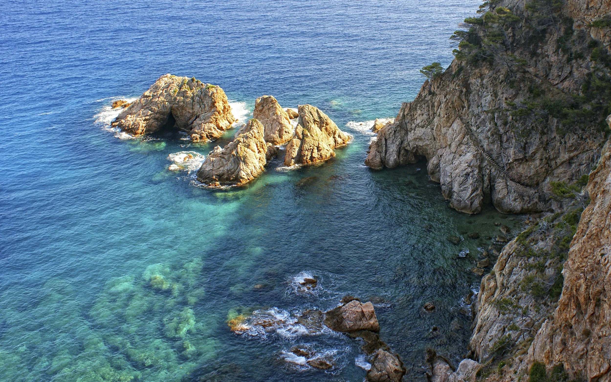 A coastal scene with turquoise water, several boats anchored near rocky shoreline, and a rocky hillside covered with green trees under a clear blue sky.