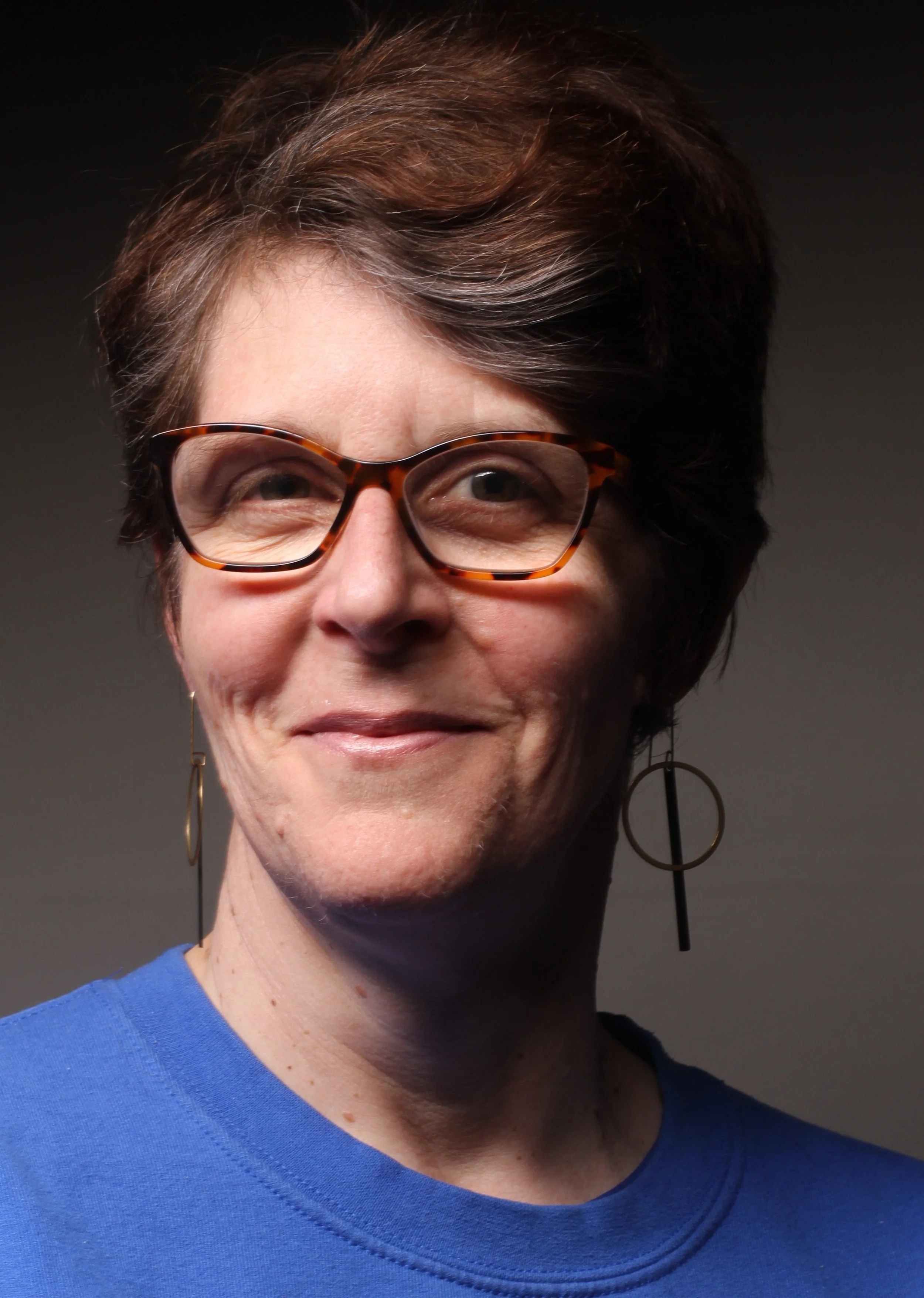 Close-up of a middle-aged white woman with short brown hair, tortoiseshell glasses, earrings, and a blue shirt