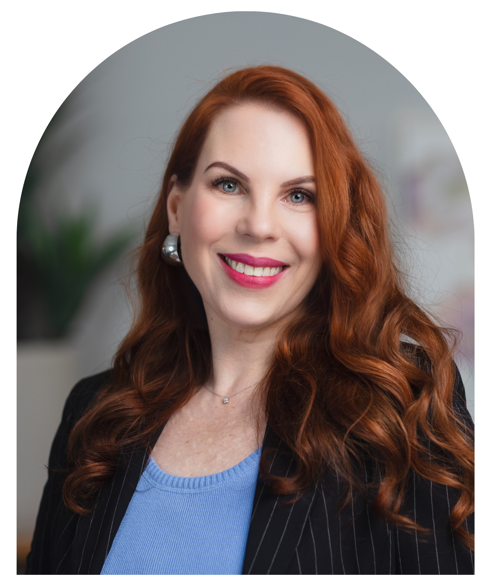 Portrait of Lyz Sutcliffe, communications advisor. She has long curly red hair, wearing a black pinstripe blazer over a light blue top, smiling with pink lipstick, earrings, and a necklace, in an indoor setting.