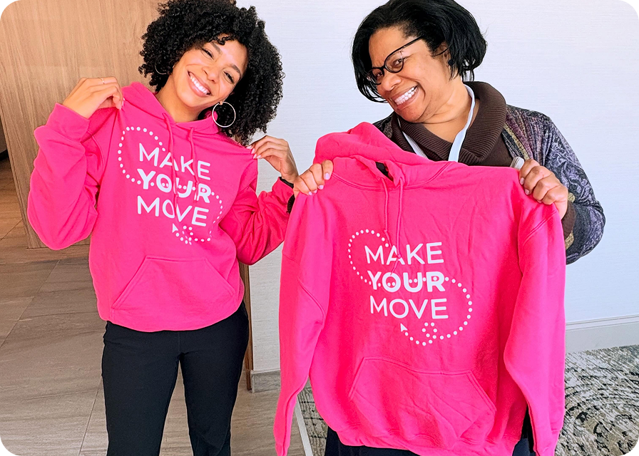 Two women holding up pink Make Your Move hoodies