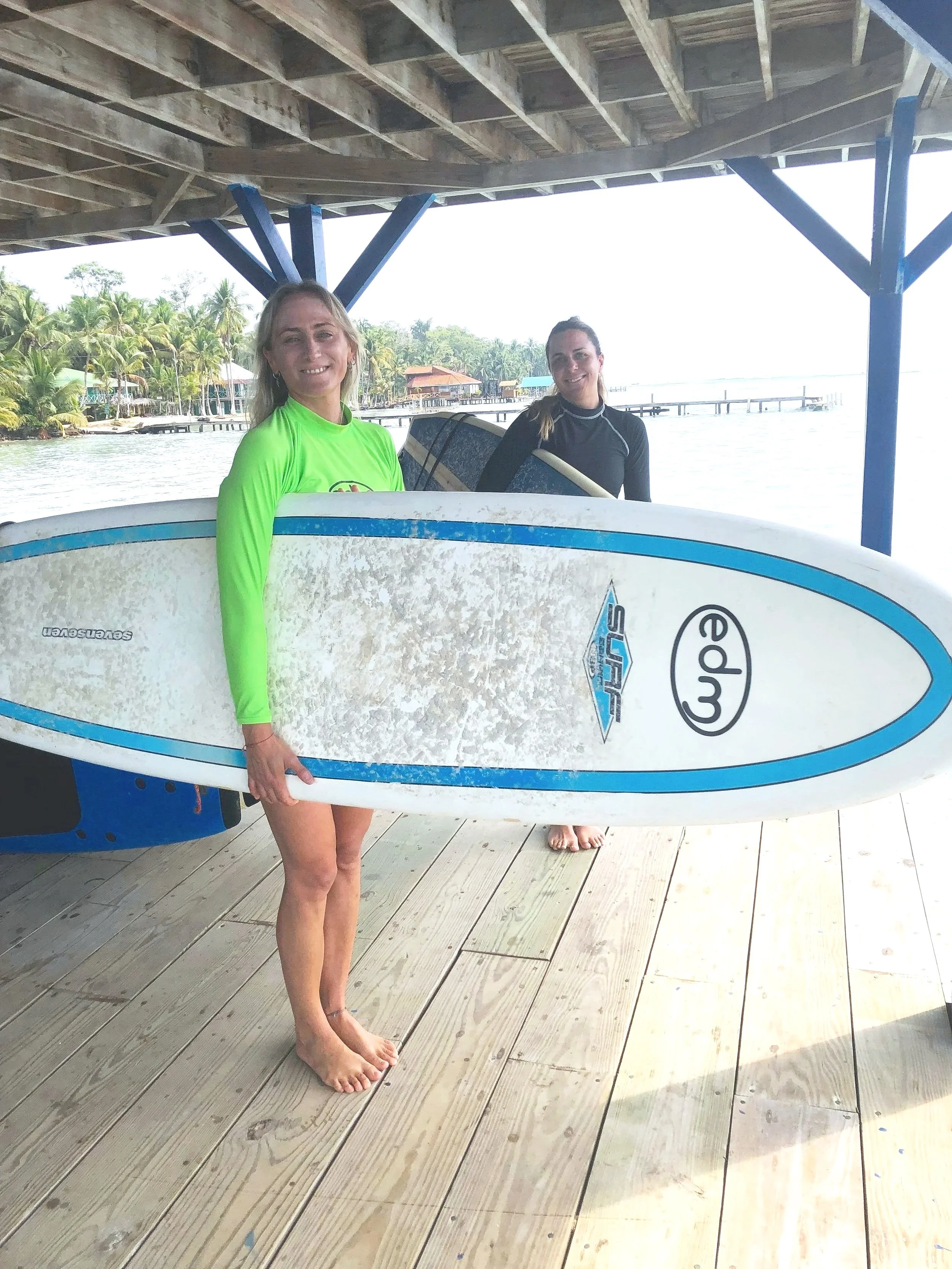 Two women holding surfboards on a wooden dock by the water, with a tropical shoreline and palm trees in the background.
