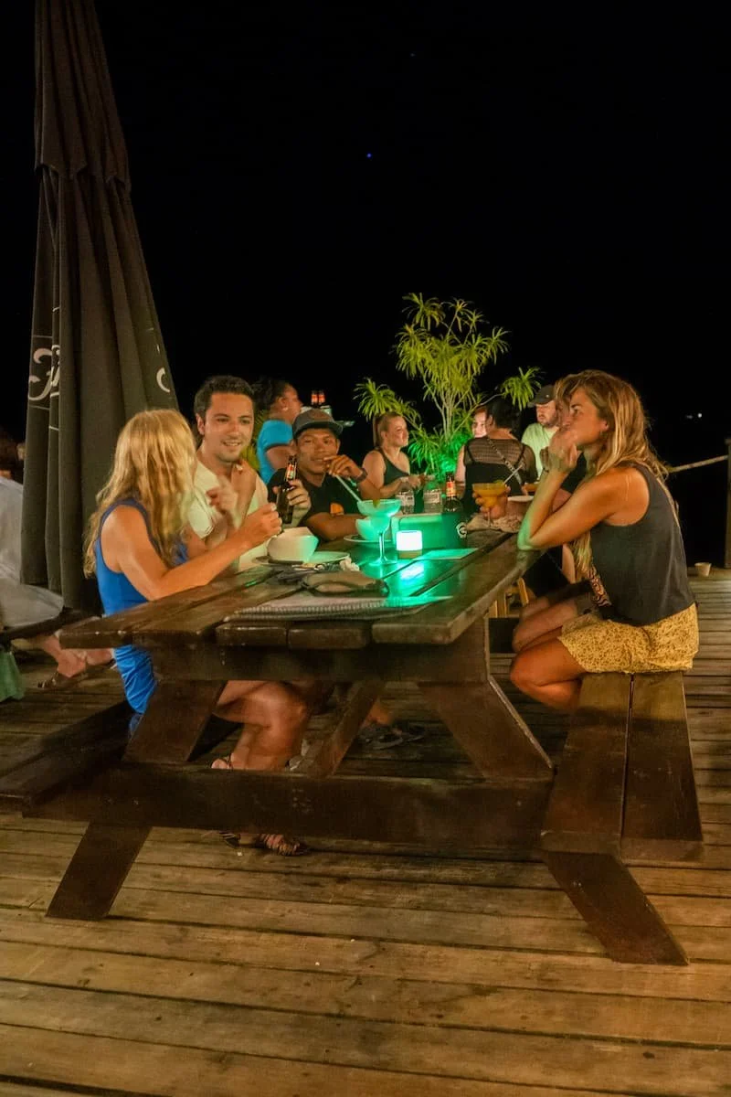A group of diverse friends enjoys a community dinner at The Experience Surf Camp in Bocas del Toro, Panama, smiling around a well-set table under warm string lights.