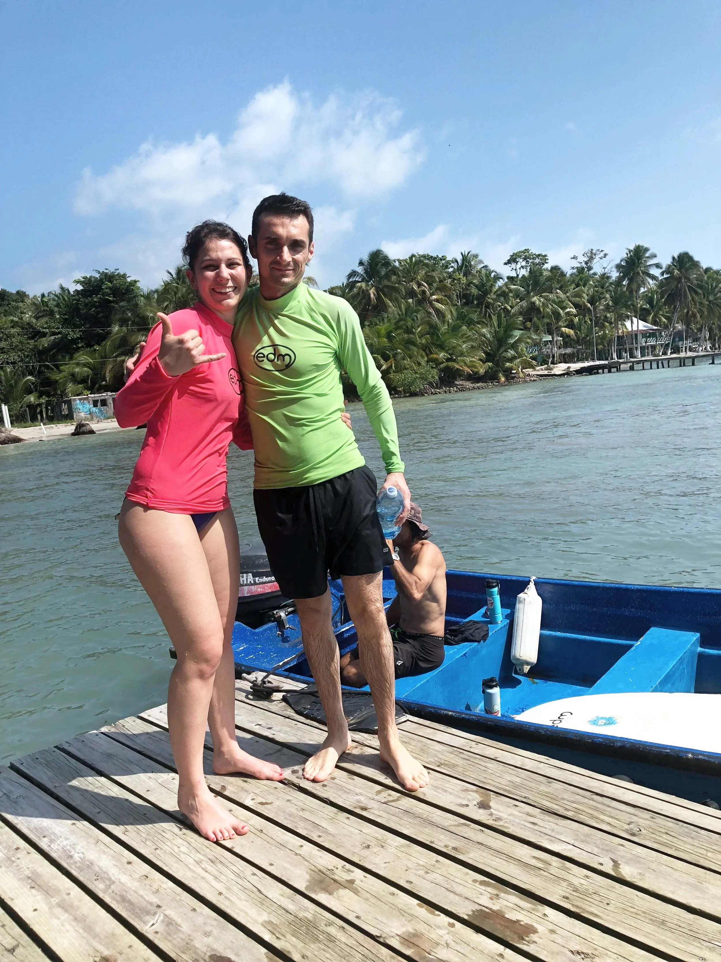 Two young adults, a woman and a man, standing on a wooden dock next to a boat by the water, smiling and posing for the camera, with palm trees and a partly cloudy sky in the background.