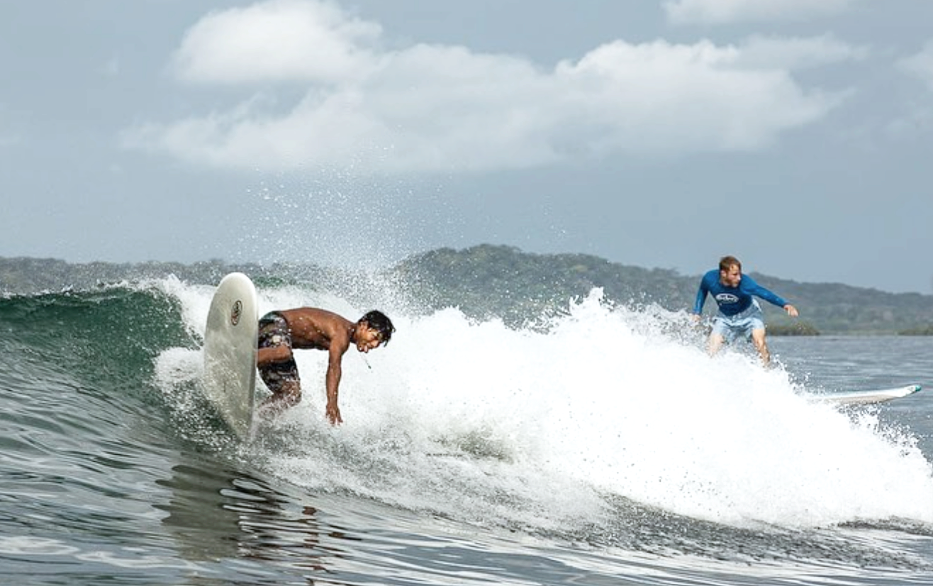 A person surfing on a wave with another person riding a surfboard in the background.