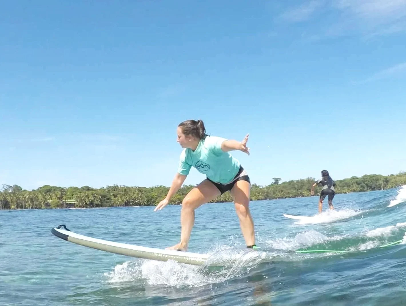 A woman in a light blue t-shirt and black shorts surfing on a paddleboard in the ocean, with another person in the background surfing as well, and a shoreline with trees in the distance under a blue sky.