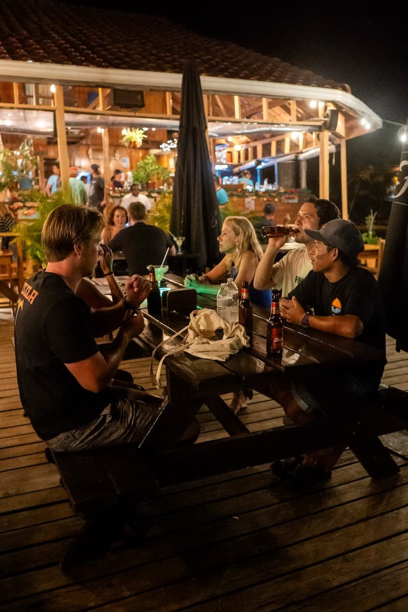 Beachside restaurant or bar on stilts at night, illuminated with string lights, reflecting on wet sand, with full moon and clear sky in the background.