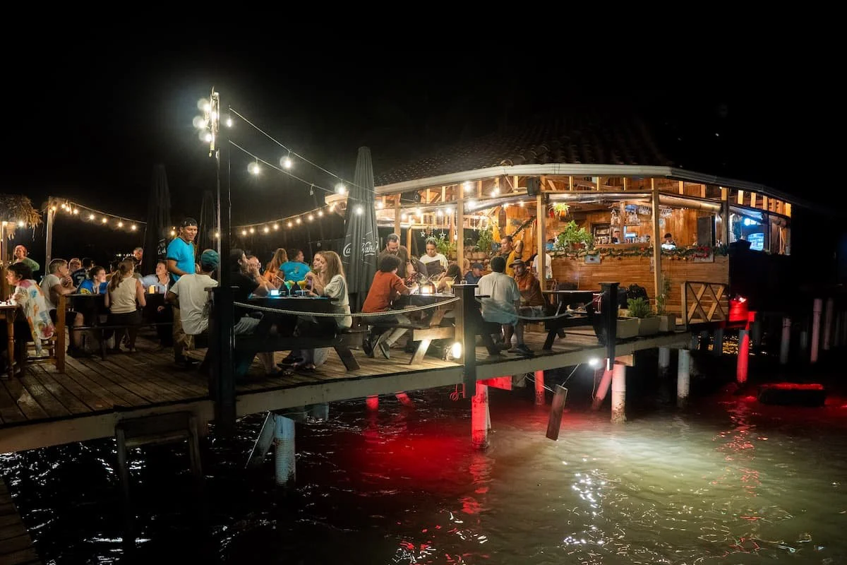 Guests enjoying drinks and dinner on the overwater wooden deck at The Experience Surf Camp in Bocas del Toro at night.