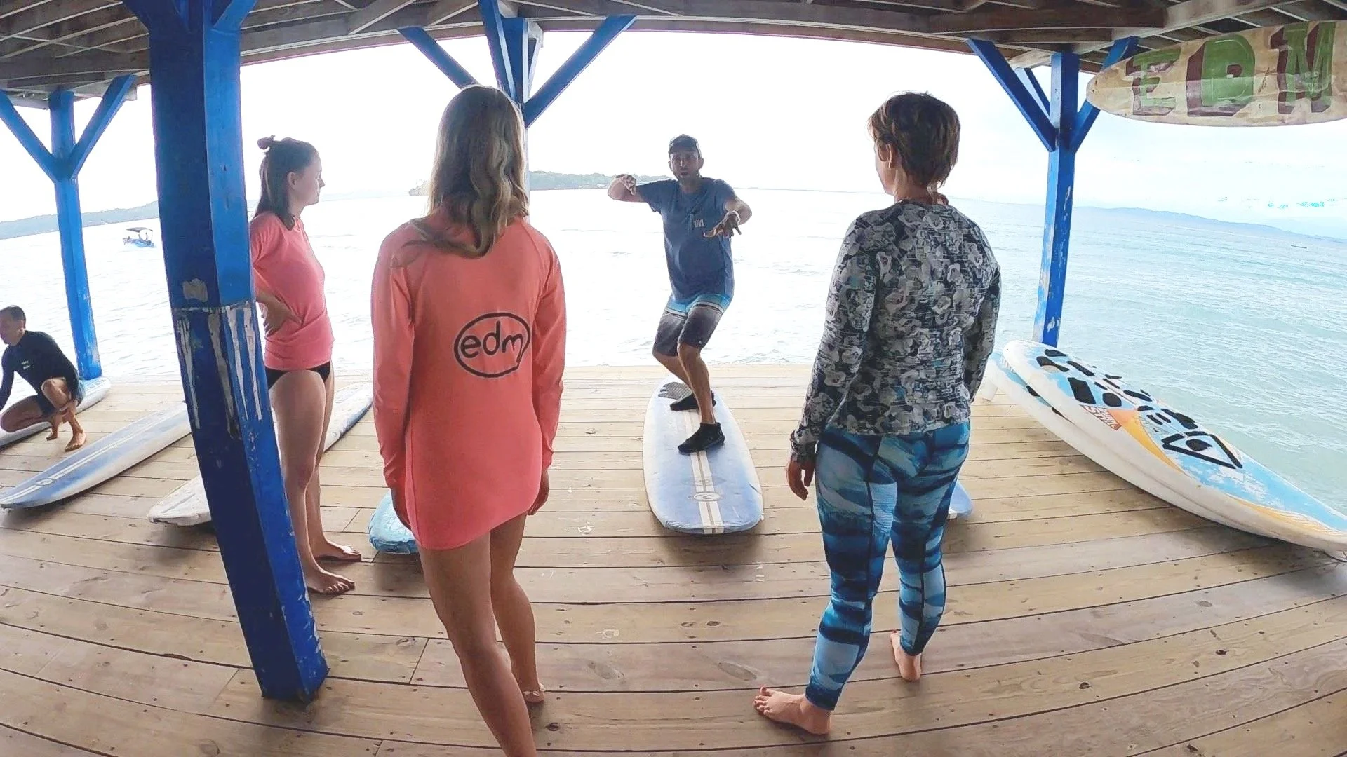 Group of people on a wooden dock by the water, with a surf instructor demonstrating balance on a surfboard, others watching, and surfboards lying on the dock.