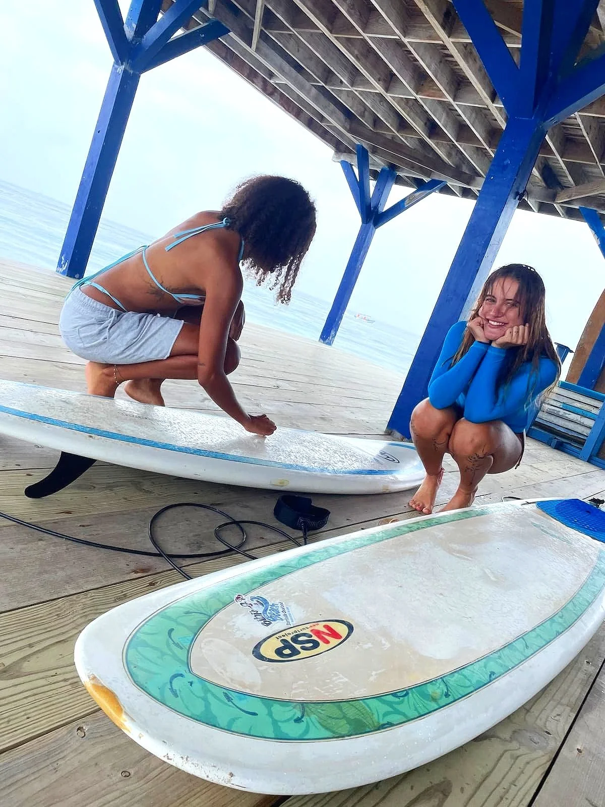 Two women on a wooden dock, one squatting and the other sitting, with surfboards nearby, under a pier at the beach.