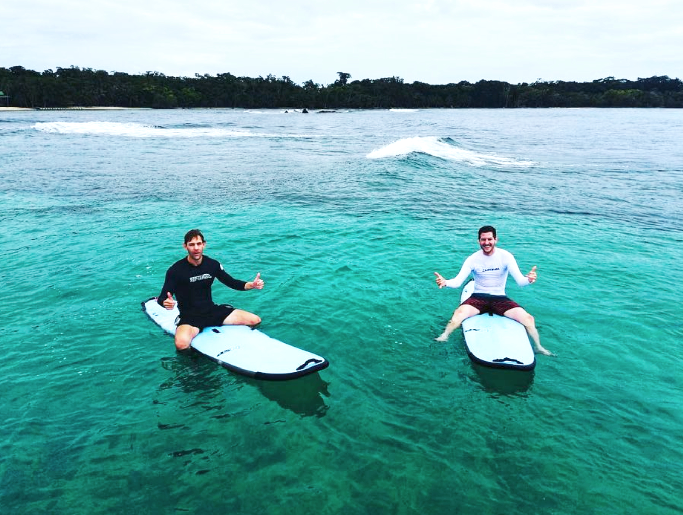 Two men sitting cross-legged on stand-up paddleboards in clear, turquoise water with a forested shoreline in the background.