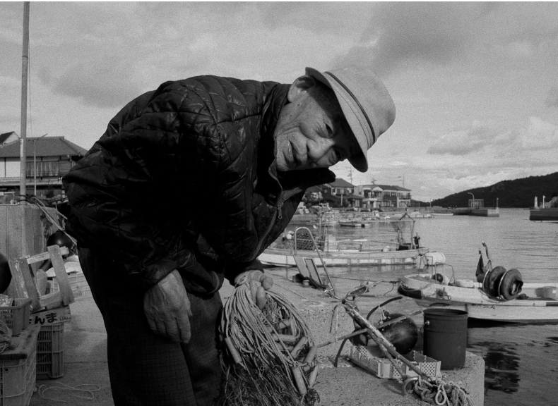 An elderly man in a quilted jacket and cap examining tangled fishing nets near a harbor with boats and houses in the background.